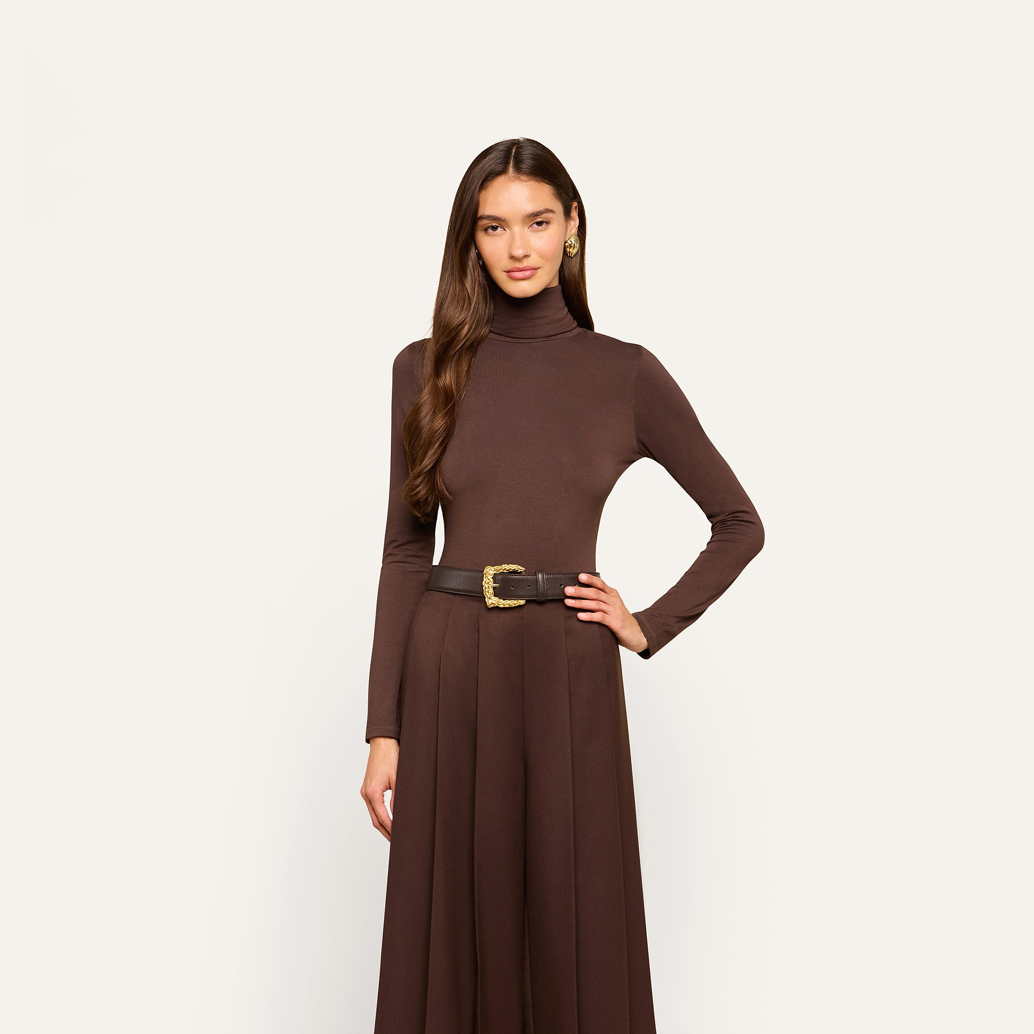 A woman stands against a plain white background wearing the Lani Turtleneck, a slim fit dress in dark java brown, styled with a wide black belt featuring a gold buckle.