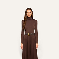 A woman stands facing forward, wearing the slim fit Lani Turtleneck in dark java brown, paired with a matching skirt and a black belt featuring a gold buckle. Her long brown hair is styled in loose waves.