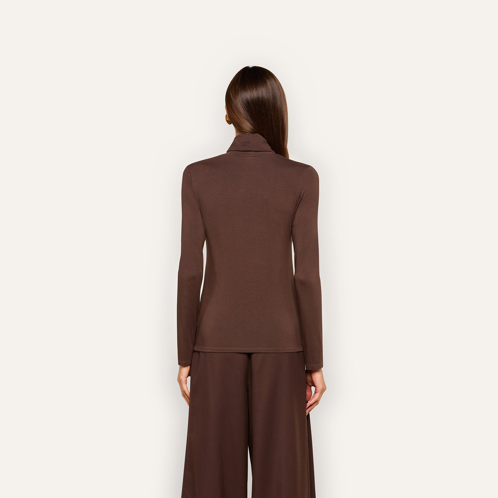 A woman with straight brown hair stands facing away, wearing the slim fit Lani Turtleneck in dark java brown and wide-leg brown pants against a plain, light background.