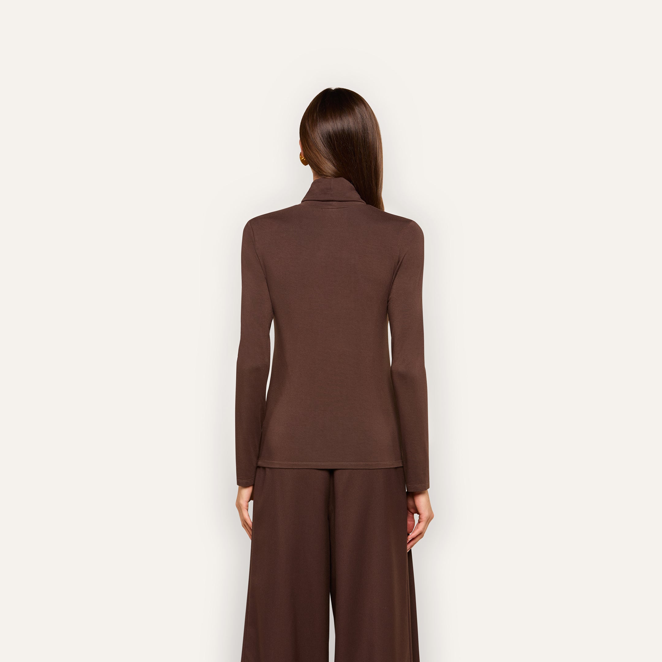 A woman with straight brown hair stands facing away, wearing the slim fit Lani Turtleneck in dark java brown and wide-leg brown pants against a plain, light background.
