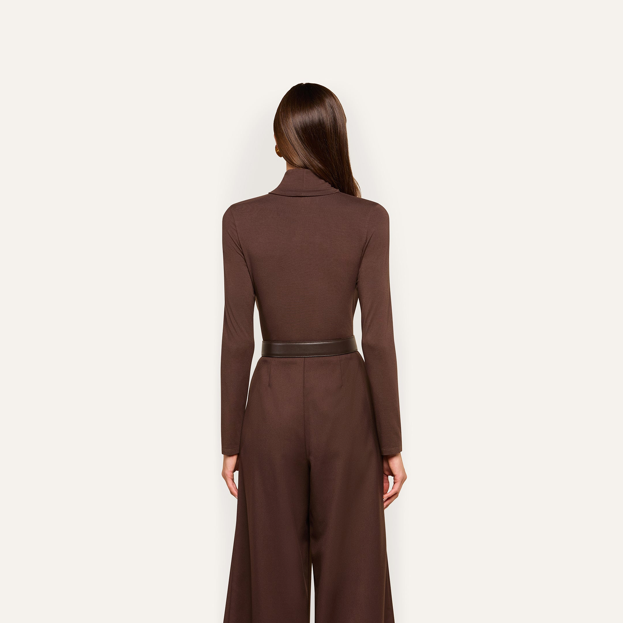 Woman standing with her back to the camera, wearing the Lani Turtleneck in dark java brown, tucked into high-waisted brown wide-leg pants, against a plain light background.
