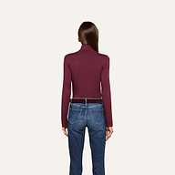 A woman stands facing away, wearing the slim fit Lani Turtleneck in maroon and blue jeans with brown belt loops, set against a plain off-white background. Perfect for Fall 2026.