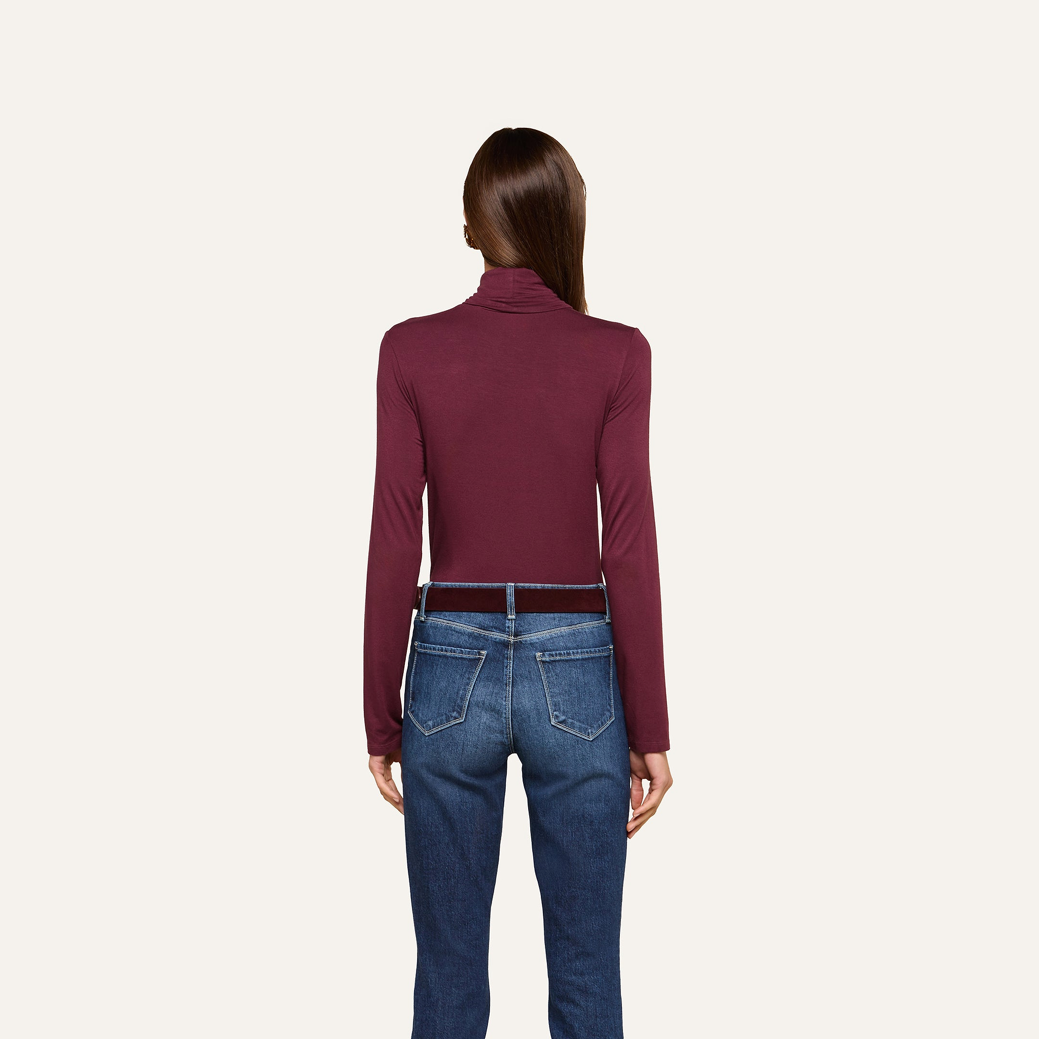 A woman stands facing away, wearing the slim fit Lani Turtleneck in maroon and blue jeans with brown belt loops, set against a plain off-white background. Perfect for Fall 2026.