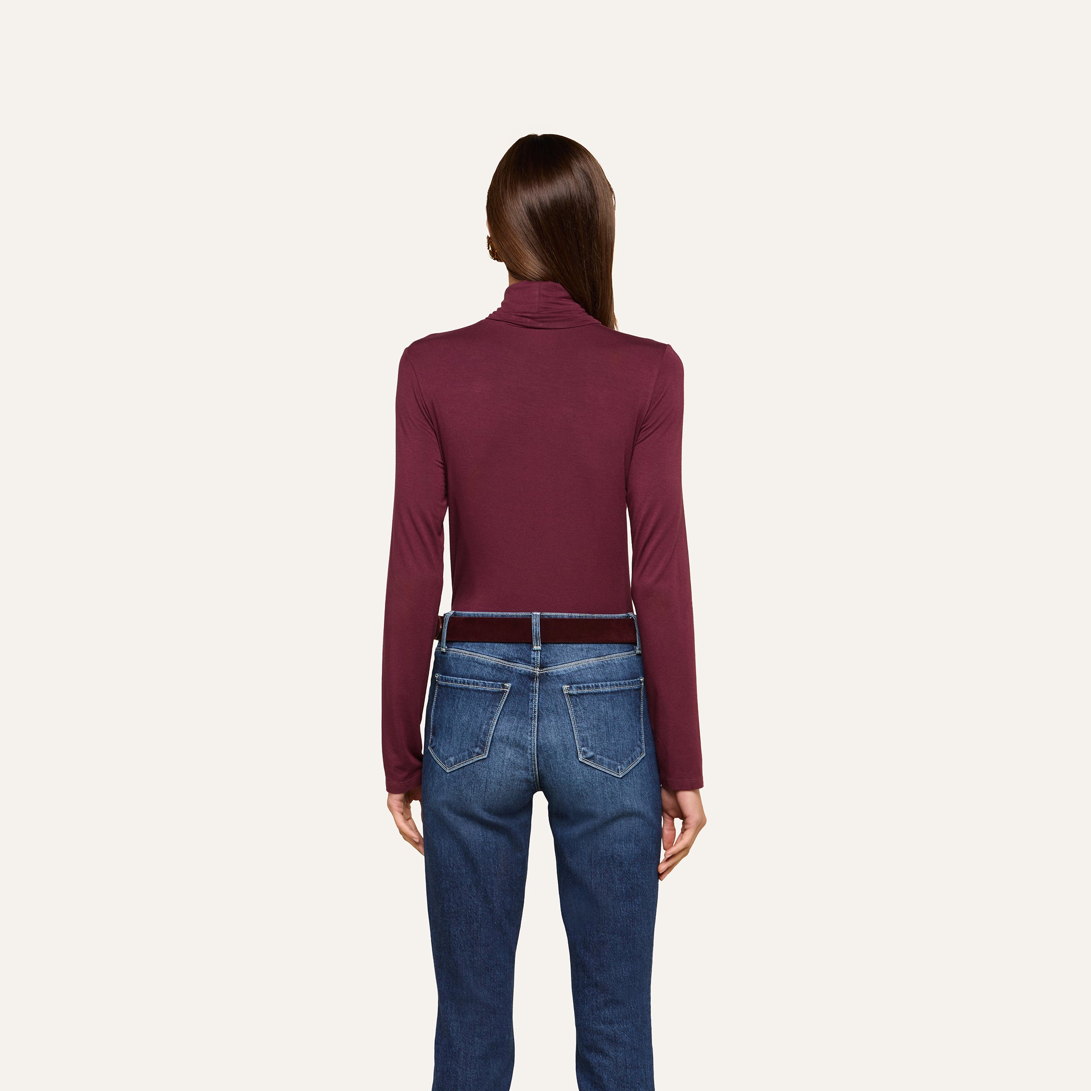 A woman stands facing away, wearing the slim fit Lani Turtleneck in maroon and blue jeans with brown belt loops, set against a plain off-white background. Perfect for Fall 2026.