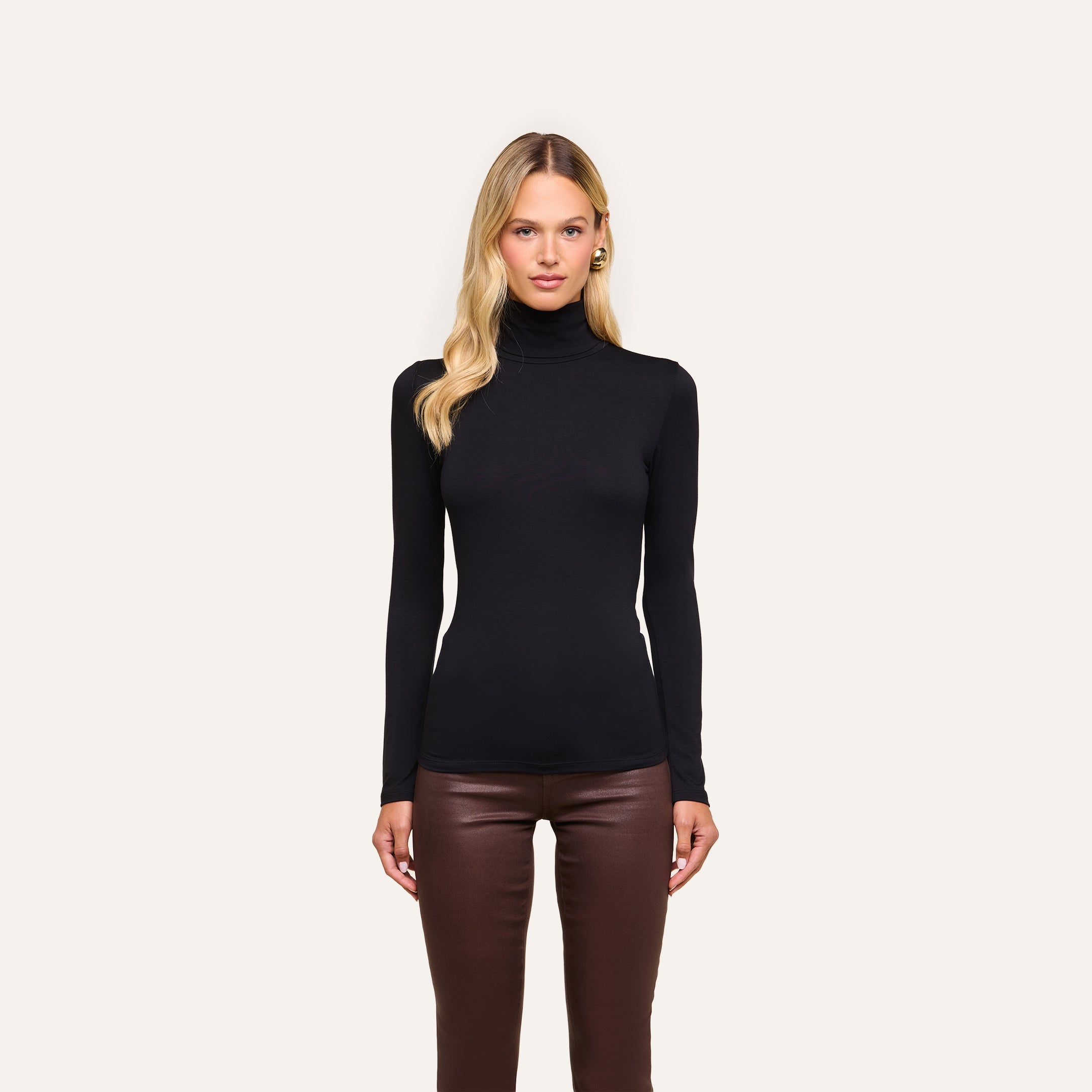 A woman stands facing forward in a slim fit Lani Turtleneck and brown leather pants against a plain light background.