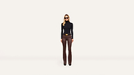 A woman in sunglasses, a black long-sleeve top, and brown pants stands confidently facing forward against a plain white background.