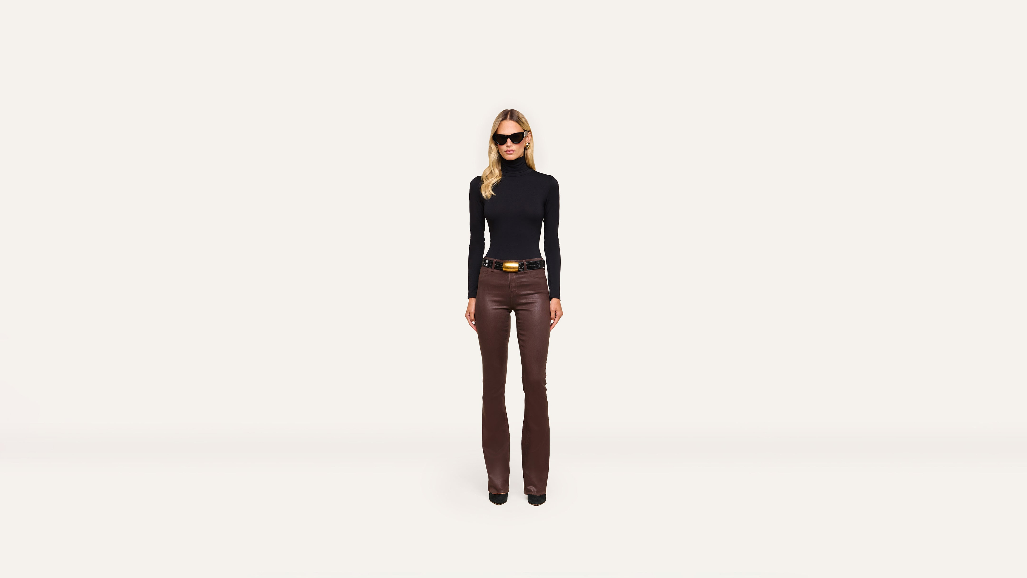 A woman in sunglasses, a black long-sleeve top, and brown pants stands confidently facing forward against a plain white background.