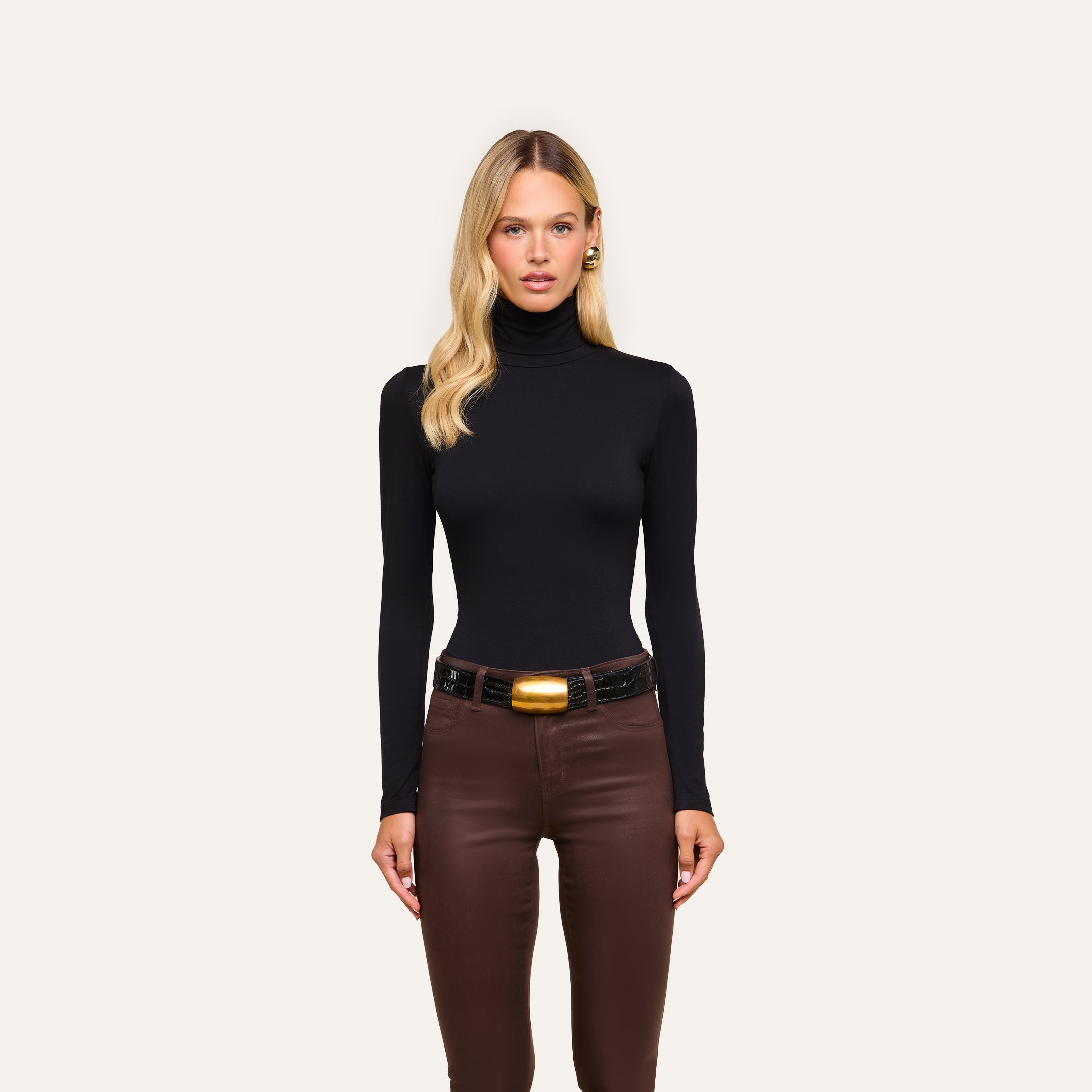 A woman with long blonde hair and a neutral expression stands facing forward in the slim fit black Lani Turtleneck, brown pants, and a black belt with a large gold buckle against a plain light background.