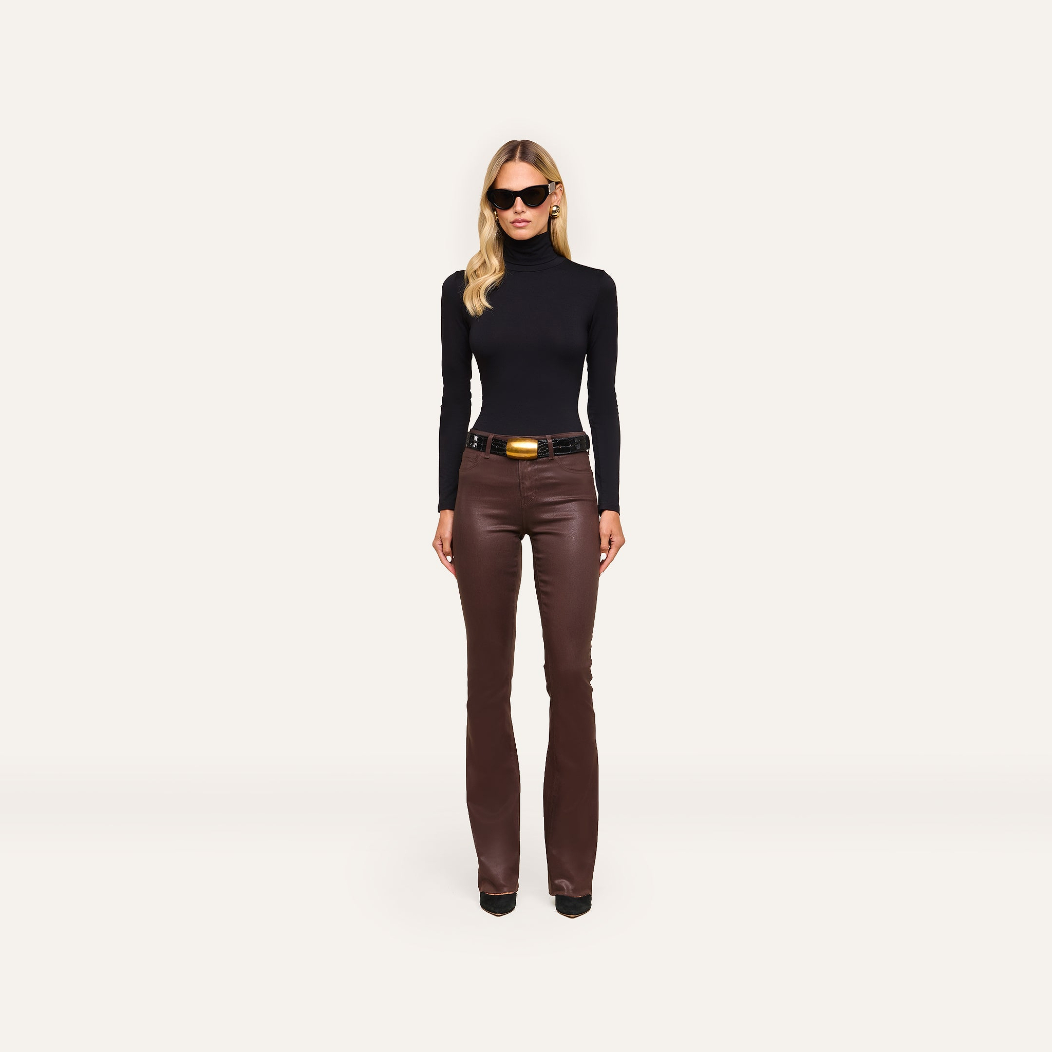 A woman in a slim fit Lani Turtleneck and brown leather pants with a belt stands against a plain white background, wearing sunglasses.