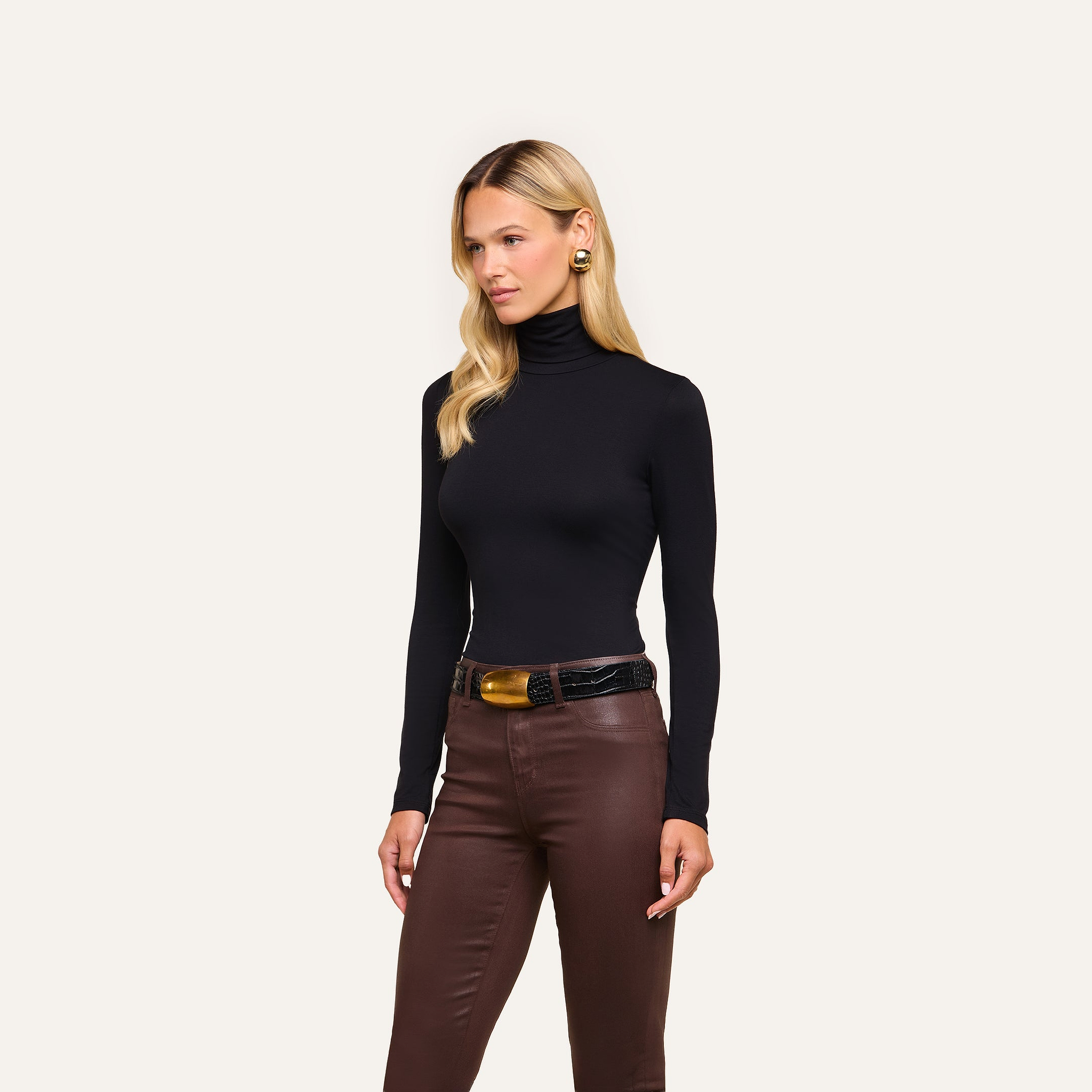 A woman with blonde hair wears the Lani Turtleneck in black, paired with brown leather pants, a black belt with a gold buckle, and gold earrings, standing against a plain light background.