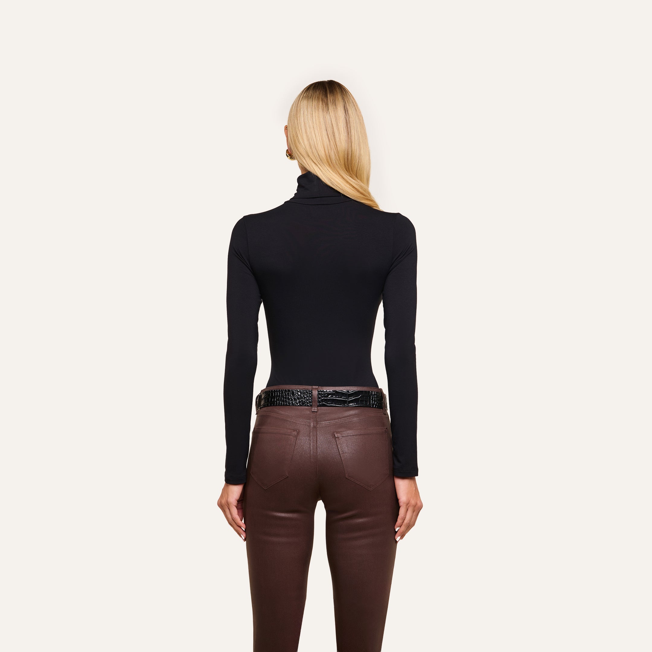 A woman with straight blonde hair models the slim fit Lani Turtleneck in black and brown pants, standing with her back to the camera against a plain white background.