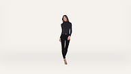 A woman in a black long-sleeve top, black pants, and heels stands against a plain white background, facing forward with a neutral expression.