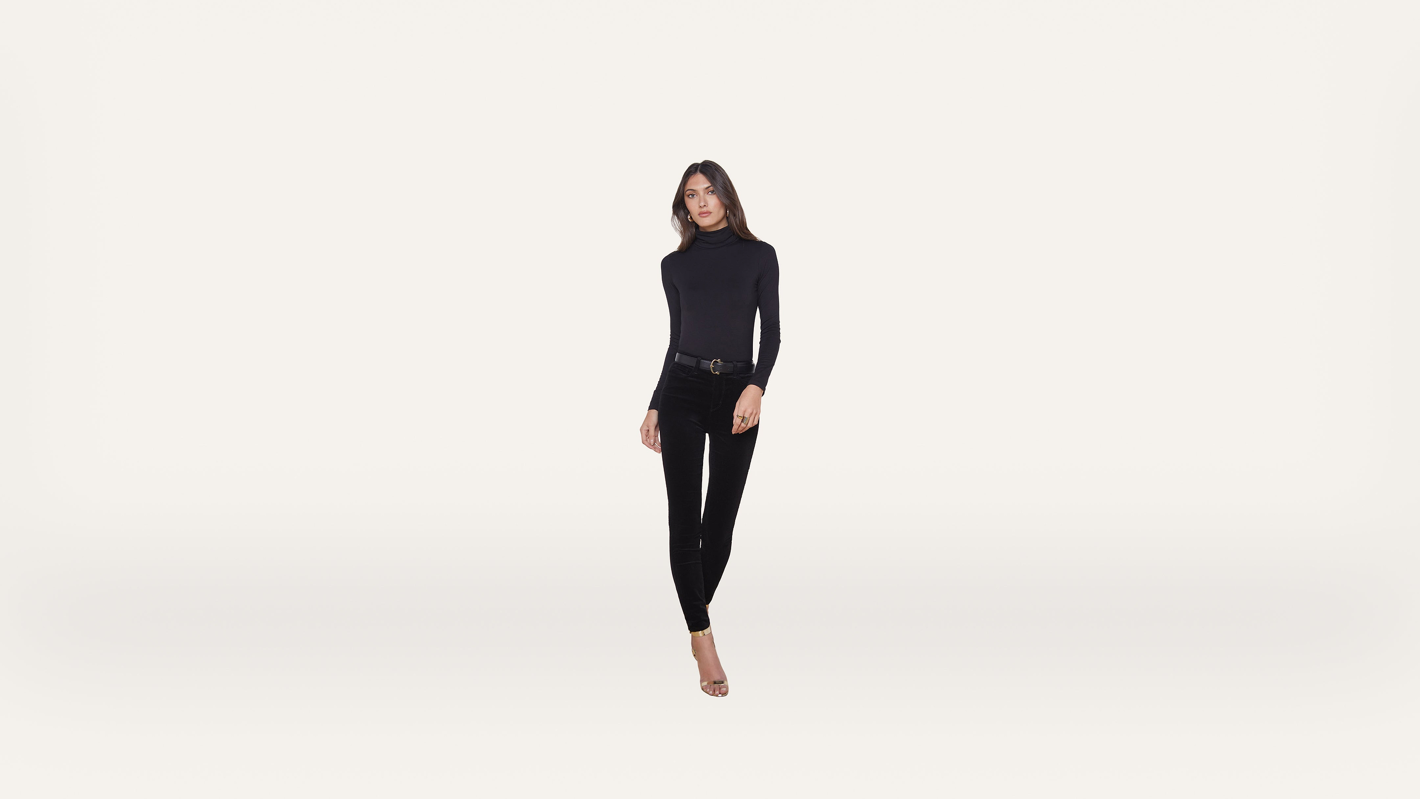 A woman in a black long-sleeve top, black pants, and heels stands against a plain white background, facing forward with a neutral expression.