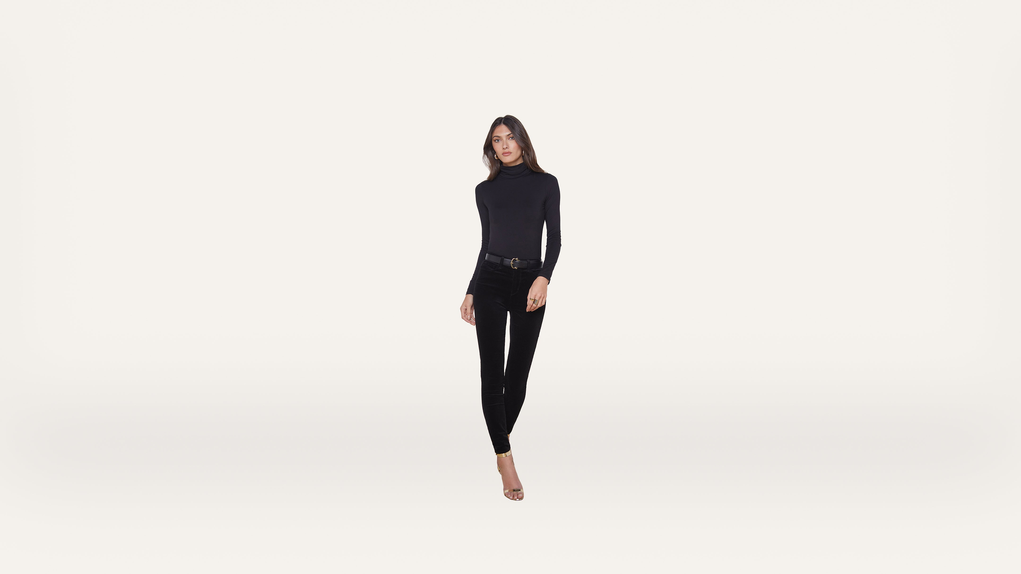 A woman in a black long-sleeve top, black pants, and heels stands against a plain white background, facing forward with a neutral expression.
