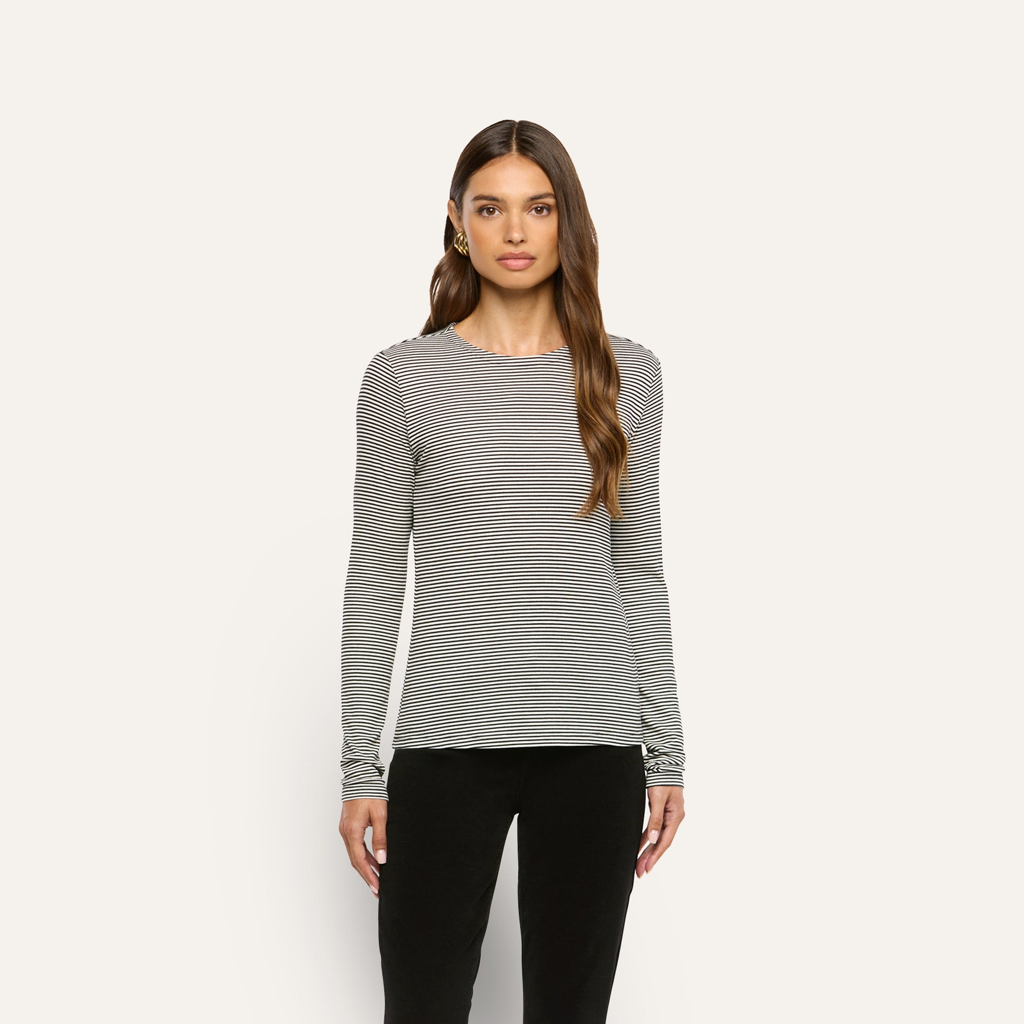 A woman wears a black-and-white thin-striped Tess Long Sleeve Tee with a slim-fit, crewneck design and black pants.