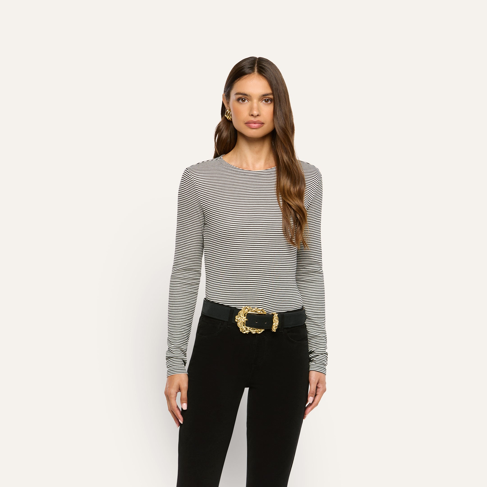 A person wearing a slim-fit, long-sleeve black and white striped Tess Long Sleeve Tee tucked into black pants with a black belt featuring a large gold buckle.
