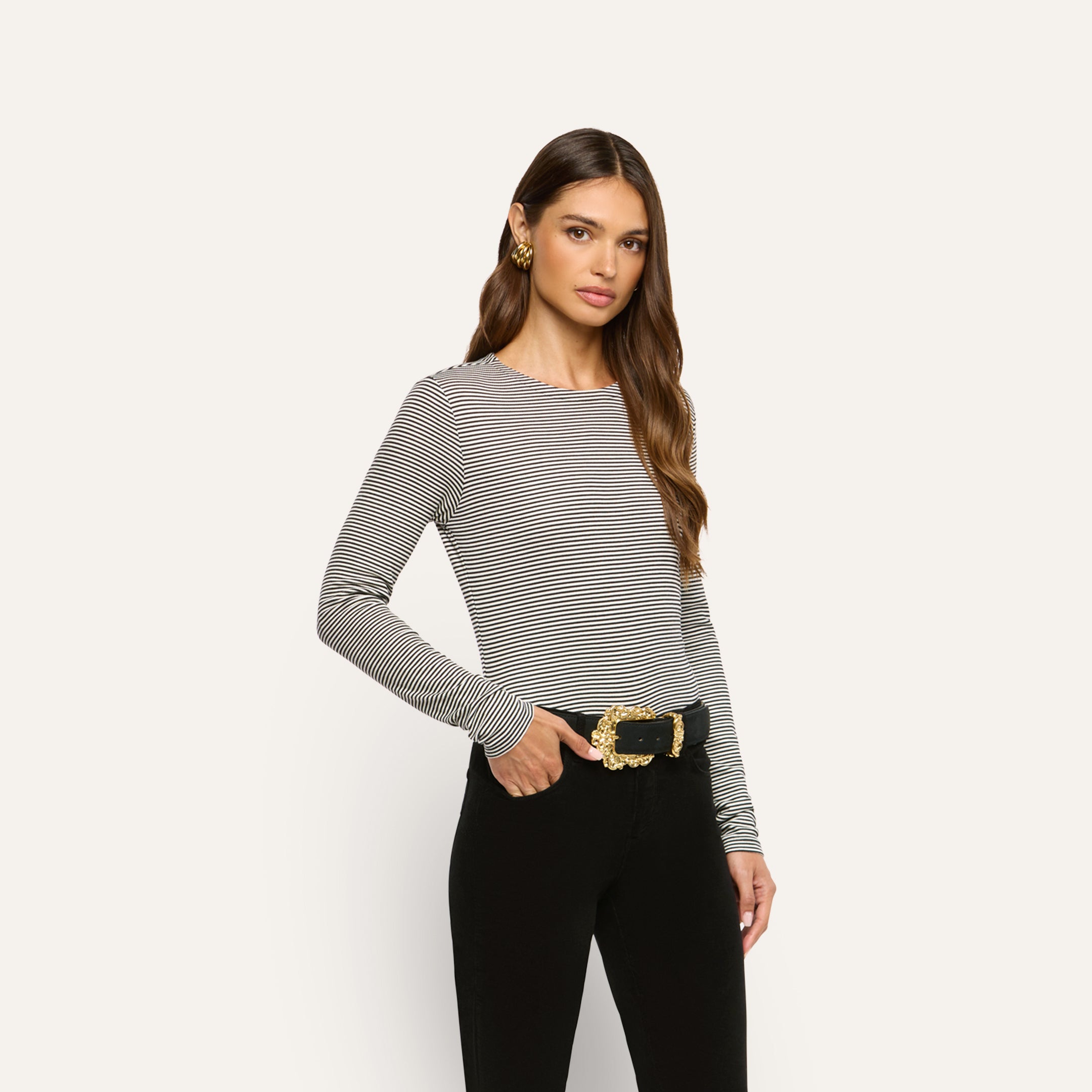 Woman wearing a slim-fit, black-and-white striped Tess Long Sleeve Tee tucked into black pants with a gold-buckled belt.