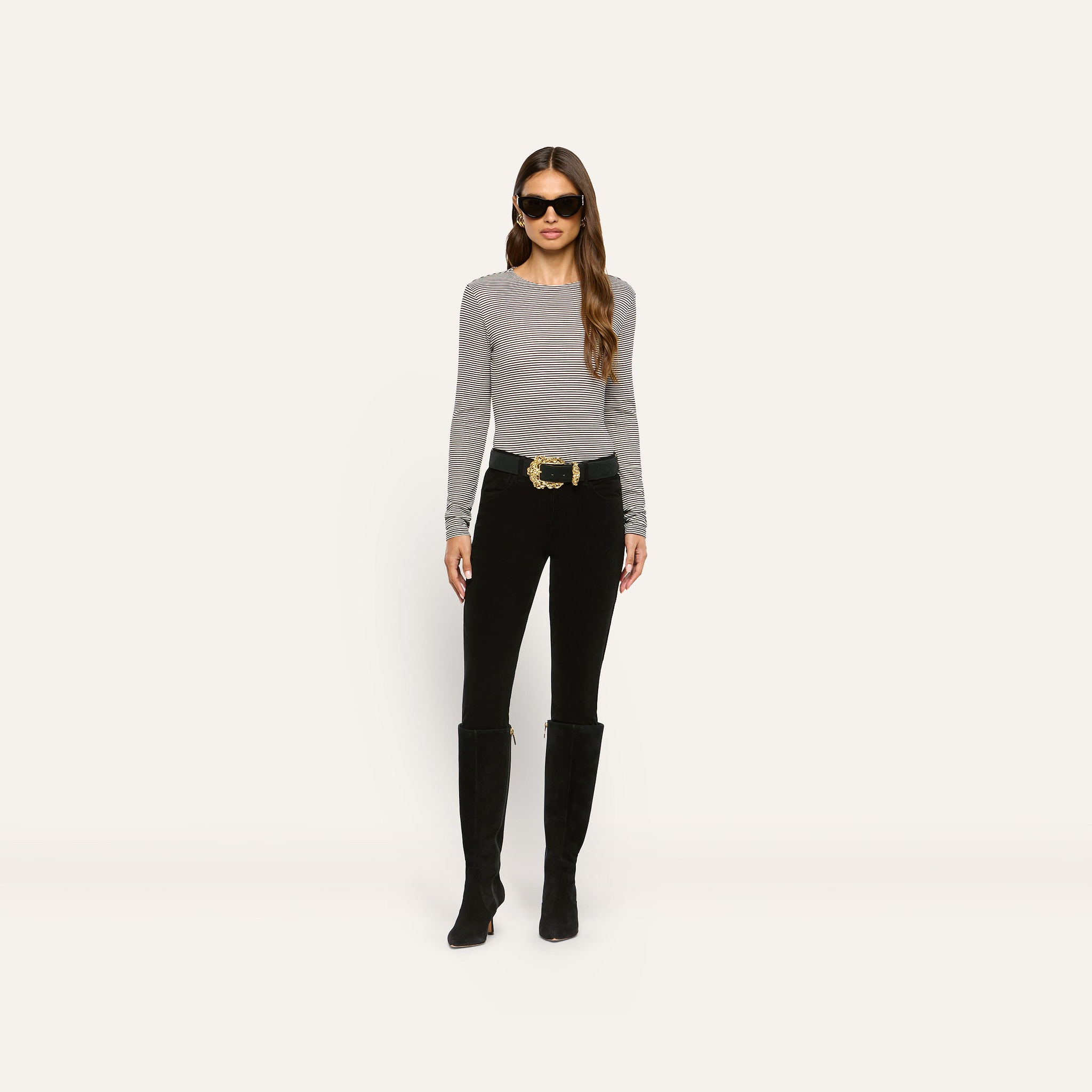 A person wearing a slim-fit, grey and white horizontal striped ribbed knit Tess Long Sleeve Tee, black pants with a gold-buckle belt, and black knee-high boots.