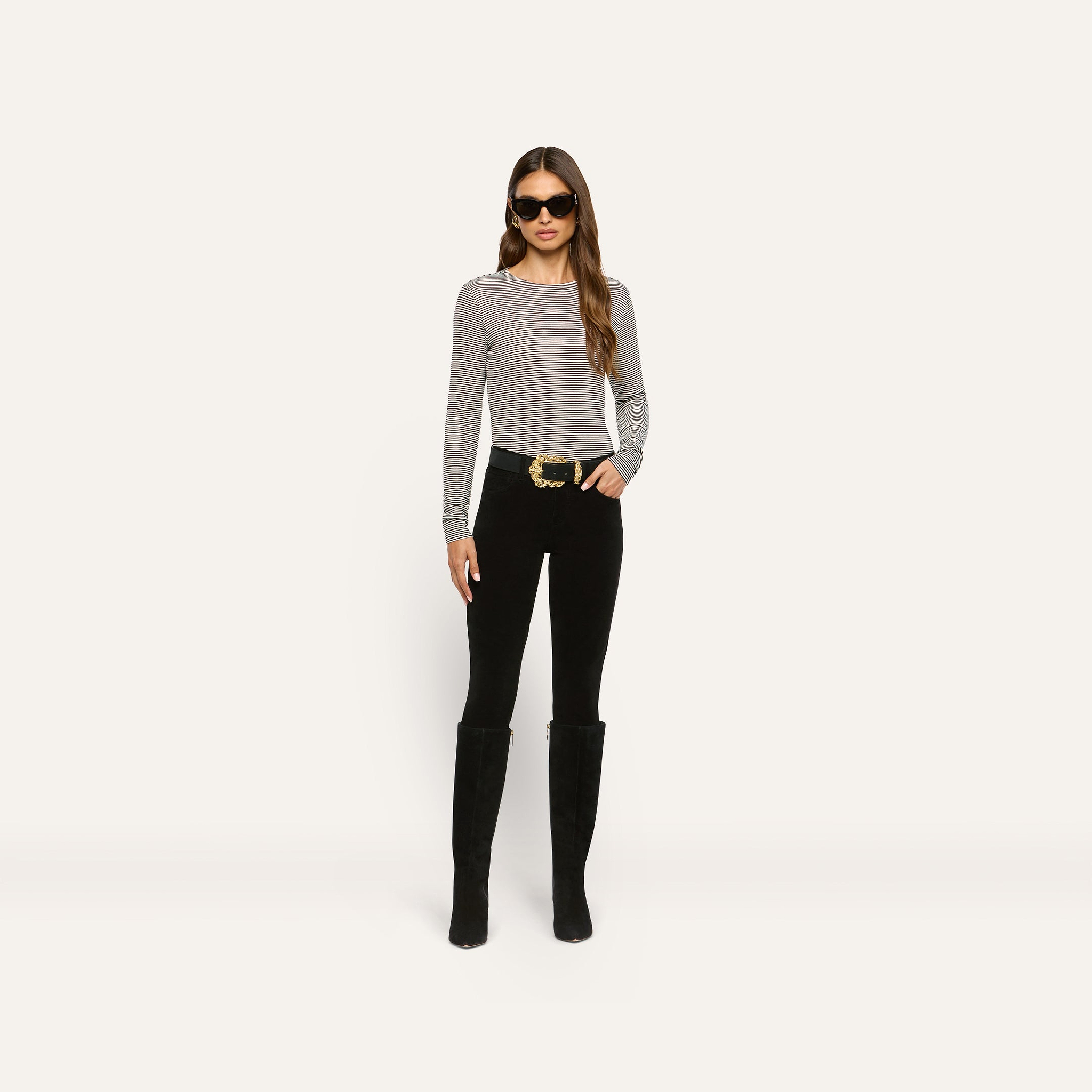 A person wearing a slim-fit, long-sleeve striped Tess Long Sleeve Tee, black pants with a large gold-buckle belt, and tall black boots.