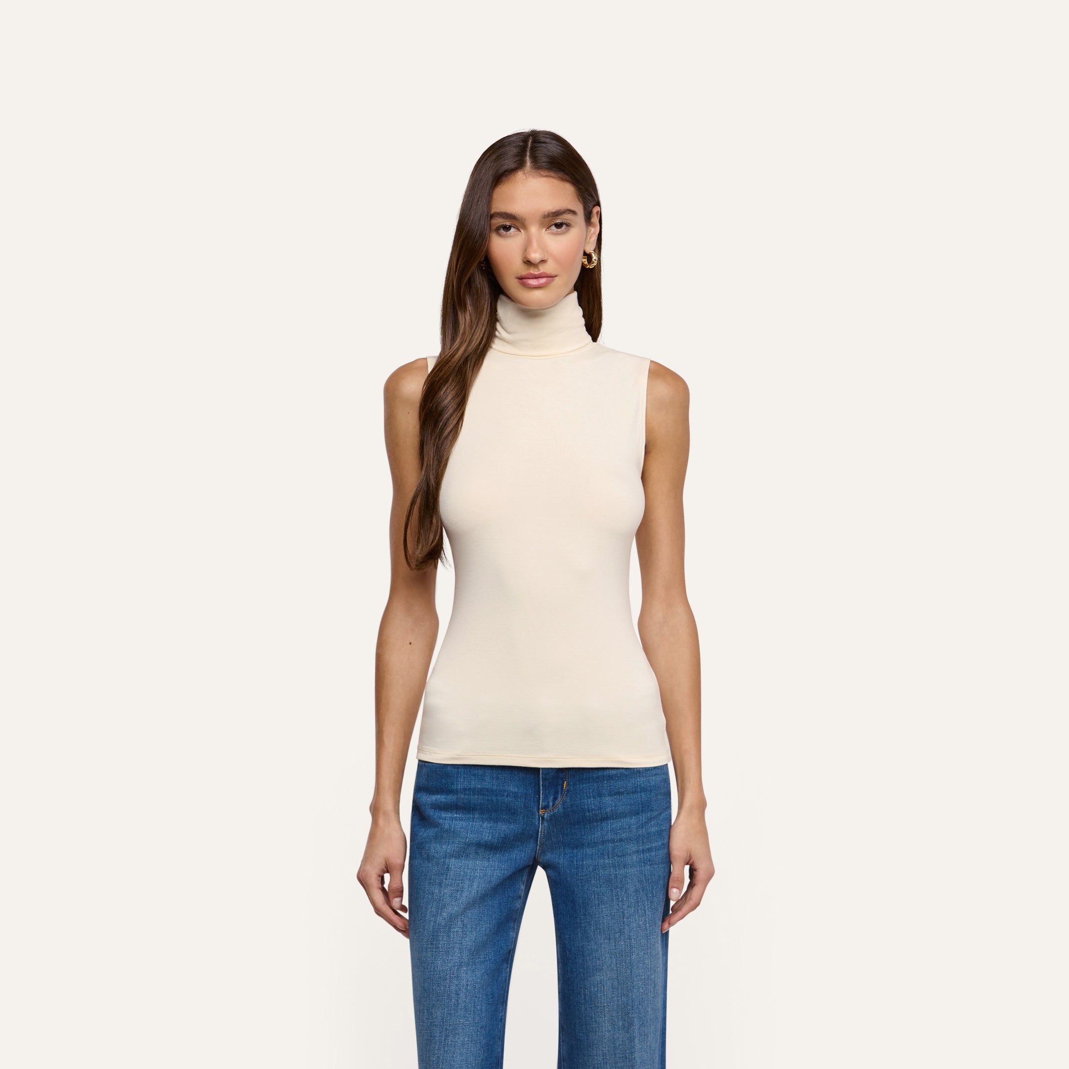 A woman wearing a cream-colored, slim-fit Ceci Sleeveless Turtleneck and blue jeans.