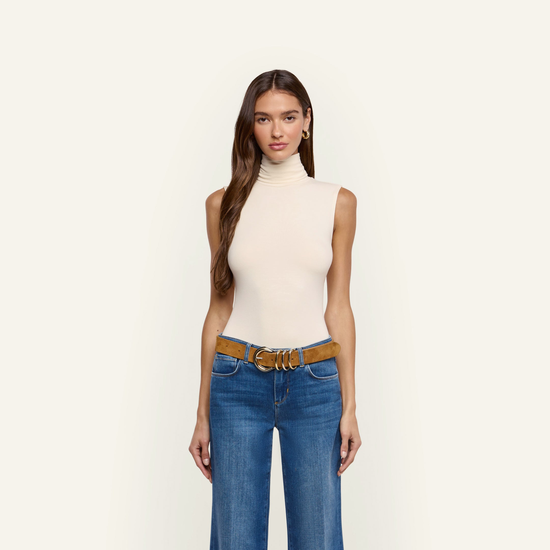 A woman wearing a cream-colored, slim-fit Ceci Sleeveless Turtleneck tucked into blue jeans with a brown suede belt.