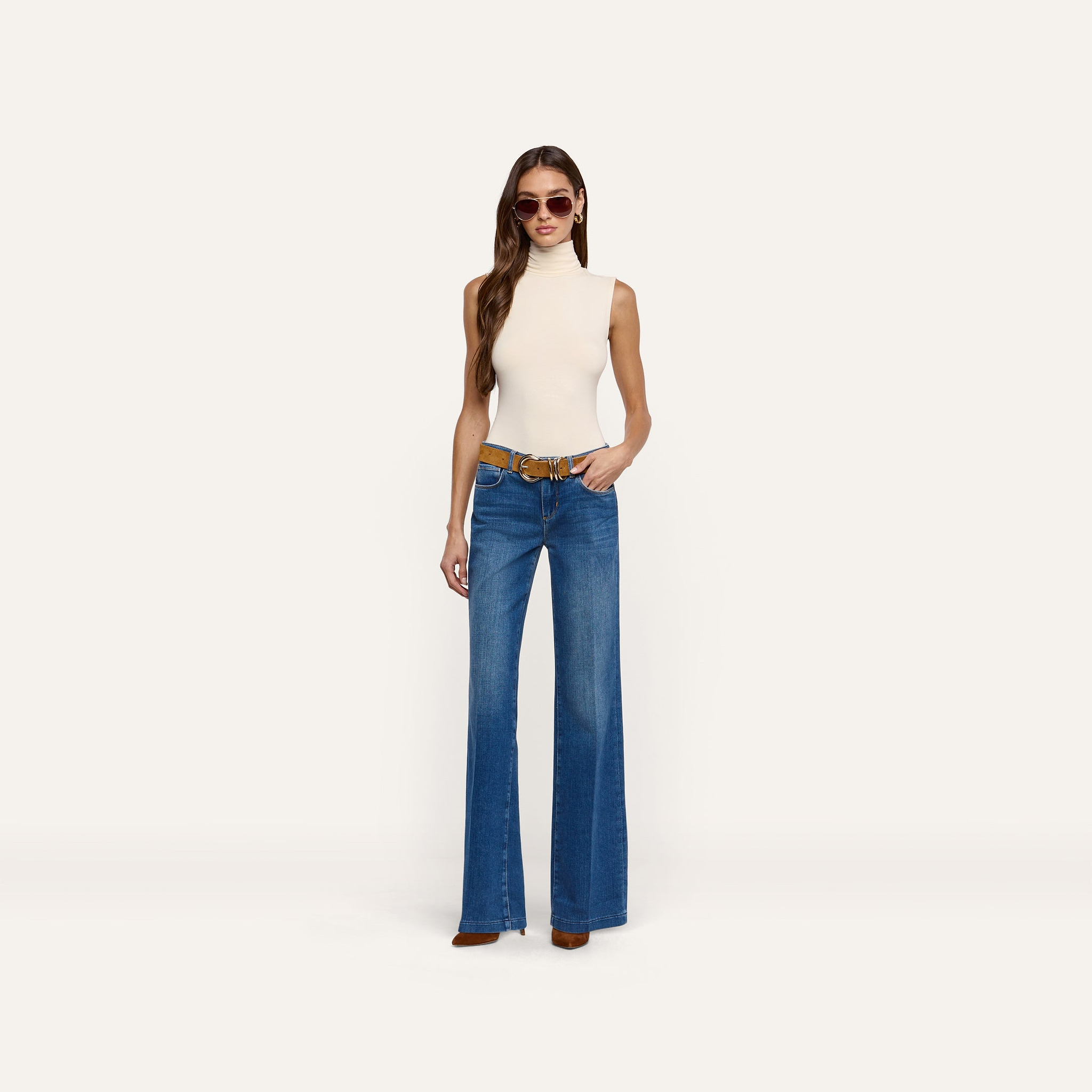 A woman wearing a cream-colored knit Ceci Sleeveless Turtleneck paired with blue wide-leg jeans and a brown belt, a timeless wardrobe staple.