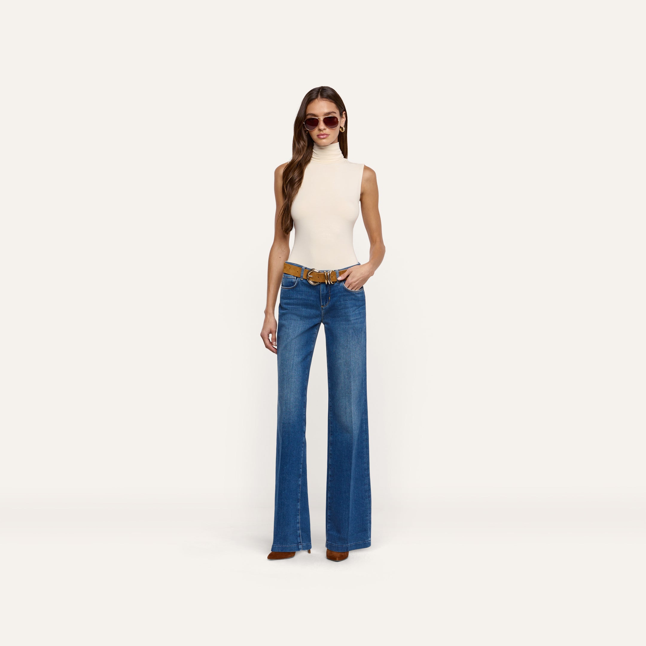 A woman wearing a cream-colored knit Ceci Sleeveless Turtleneck paired with blue wide-leg jeans and a brown belt, a timeless wardrobe staple.