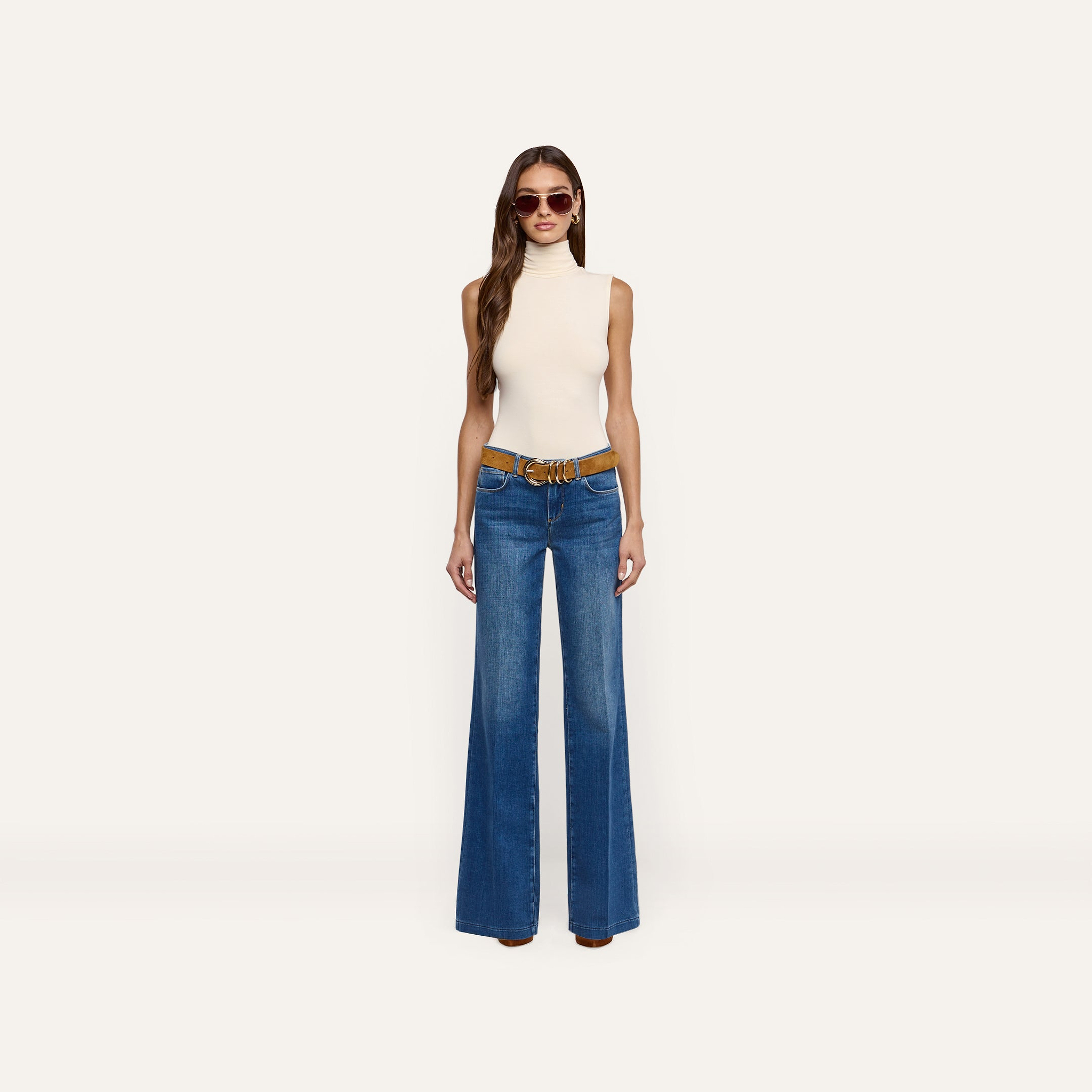 A woman wearing a cream Ceci Sleeveless Turtleneck paired with a brown belt and wide-leg blue jeans.