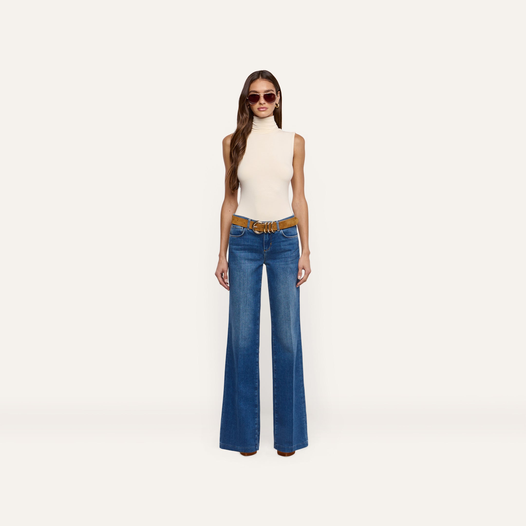 A woman wearing a cream Ceci Sleeveless Turtleneck paired with a brown belt and wide-leg blue jeans.