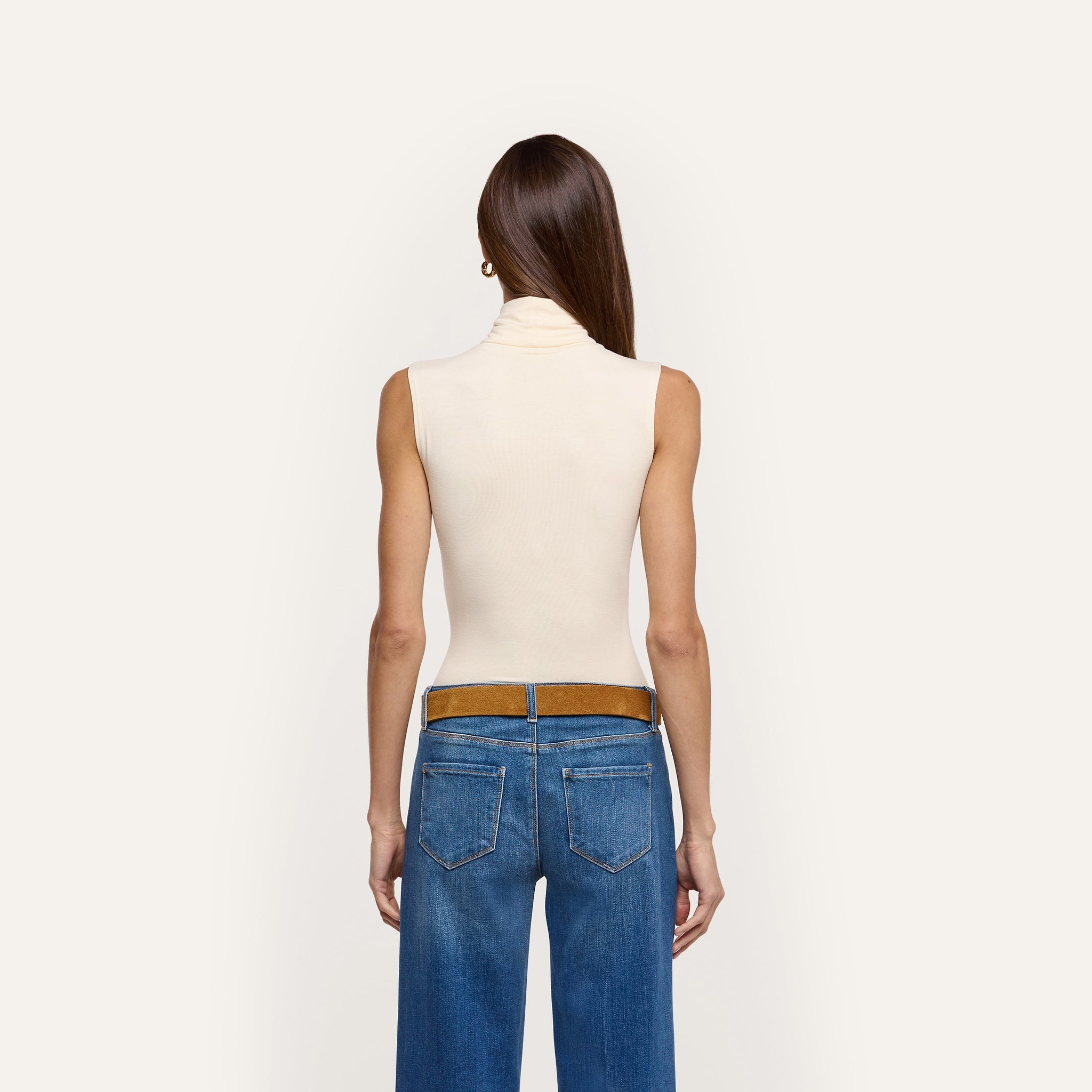 A slim-fit, sleeveless, cream-colored Ceci Sleeveless Turtleneck tucked into blue jeans with a brown belt, seen from the back.