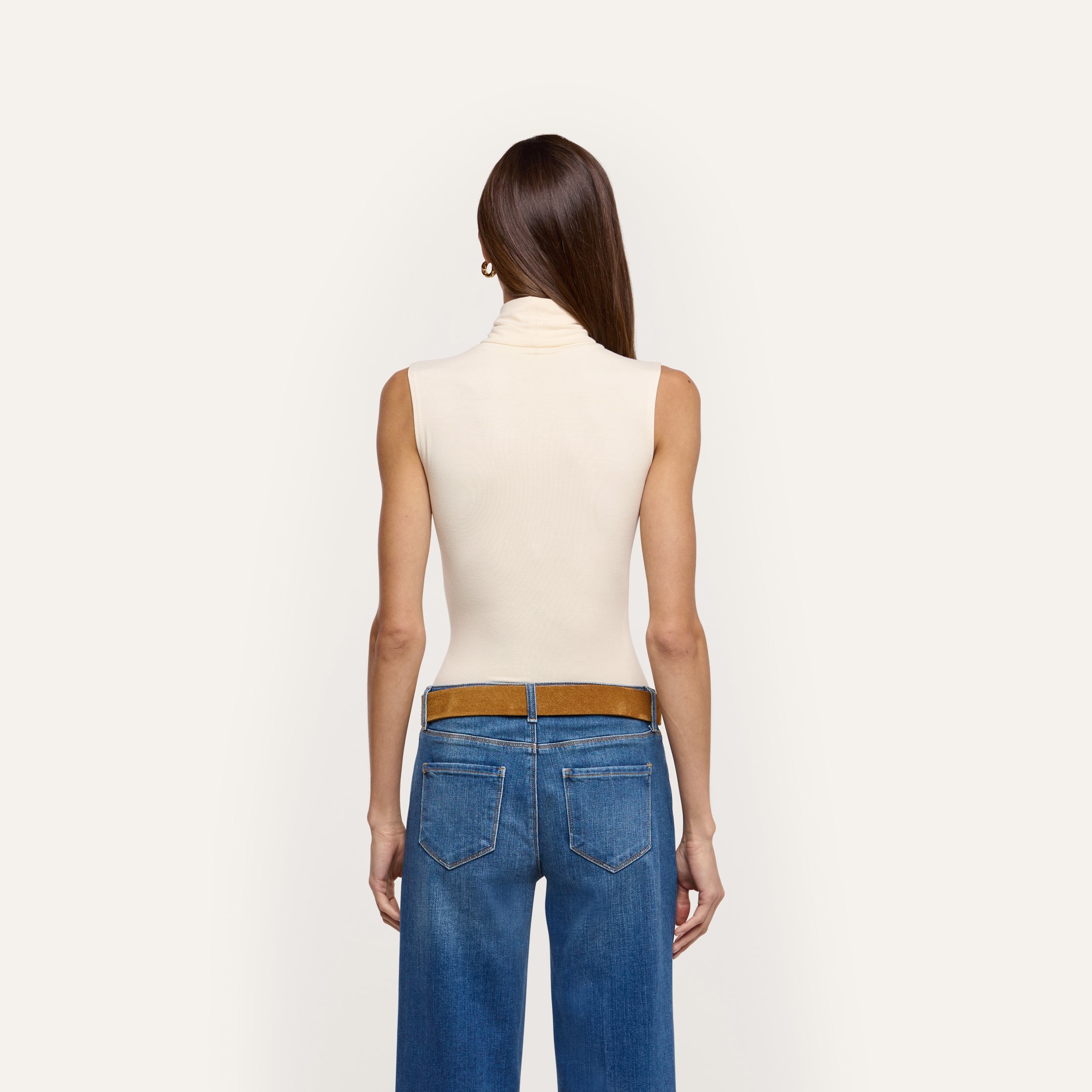 A slim-fit, sleeveless, cream-colored Ceci Sleeveless Turtleneck tucked into blue jeans with a brown belt, seen from the back.