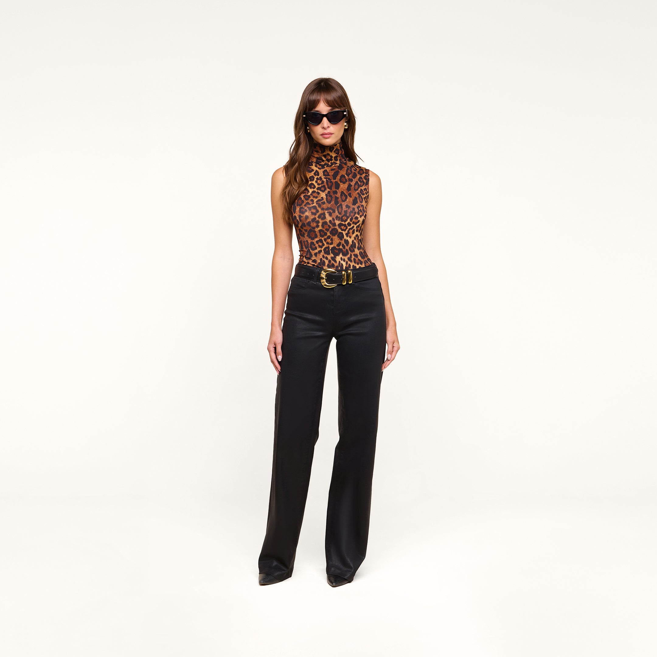 A woman wearing sunglasses, the Ceci Sleeveless Turtleneck in a leopard print, and slim fit high-waisted black pants stands against a plain white background.