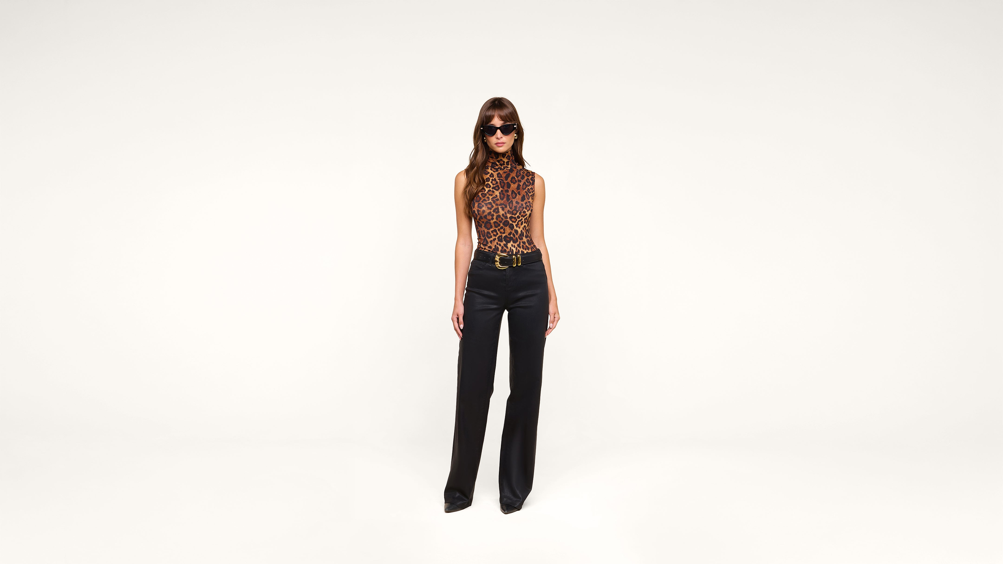 A woman wearing sunglasses, a sleeveless leopard-print top, black belted pants, and black shoes stands against a plain white background.