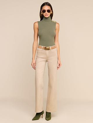 A woman in sunglasses wears the Ceci Sleeveless Turtleneck in ash olive green, styled with beige high-waisted pants featuring a wide belt and olive green boots, standing against a plain beige background.