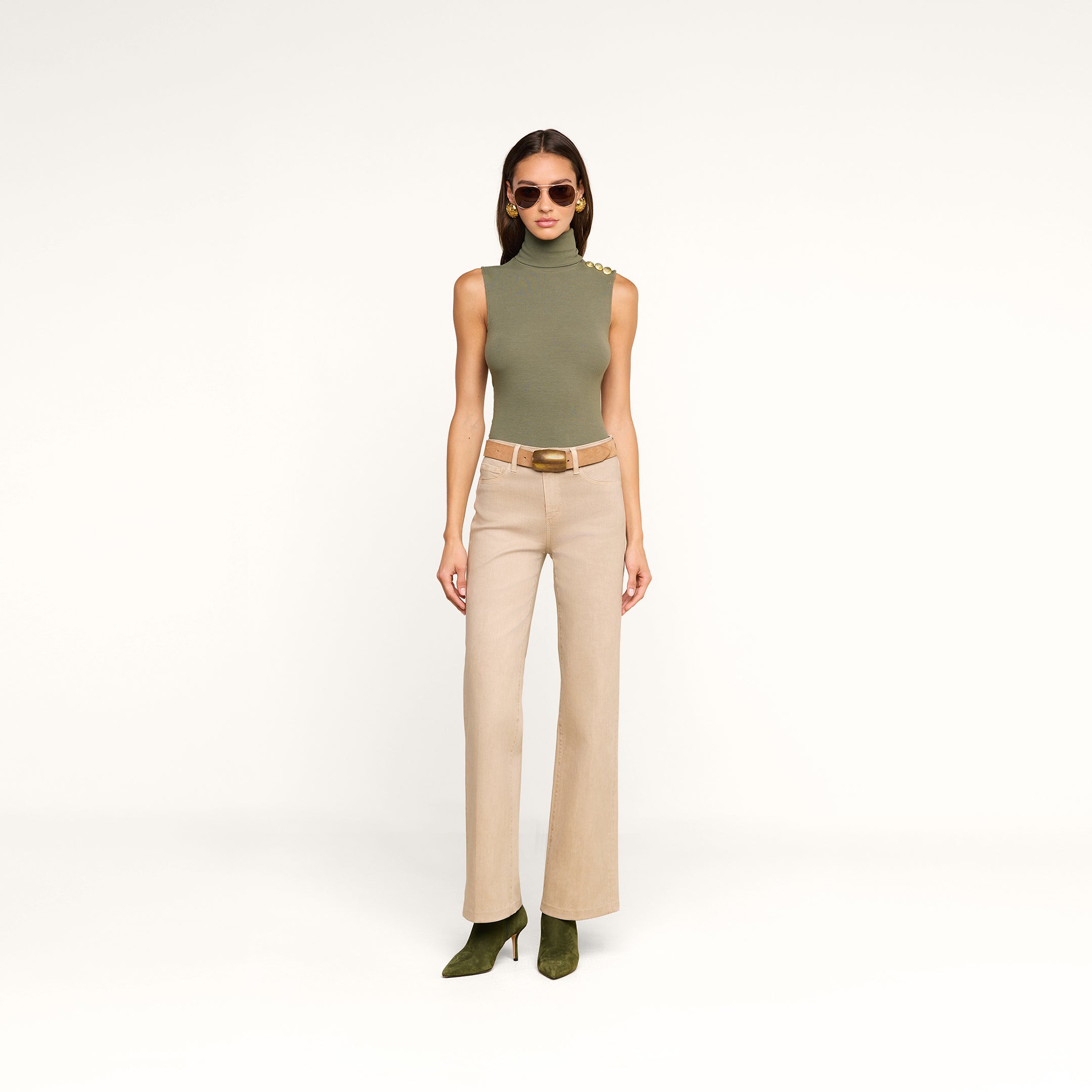 Wearing the Ceci Sleeveless Turtleneck in ash olive green, paired with beige high-waisted belted pants and green heels, a woman in sunglasses stands against a plain white background.