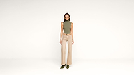 A woman wearing sunglasses, a sleeveless green turtleneck, beige high-waisted pants, and green heels stands against a plain white background.