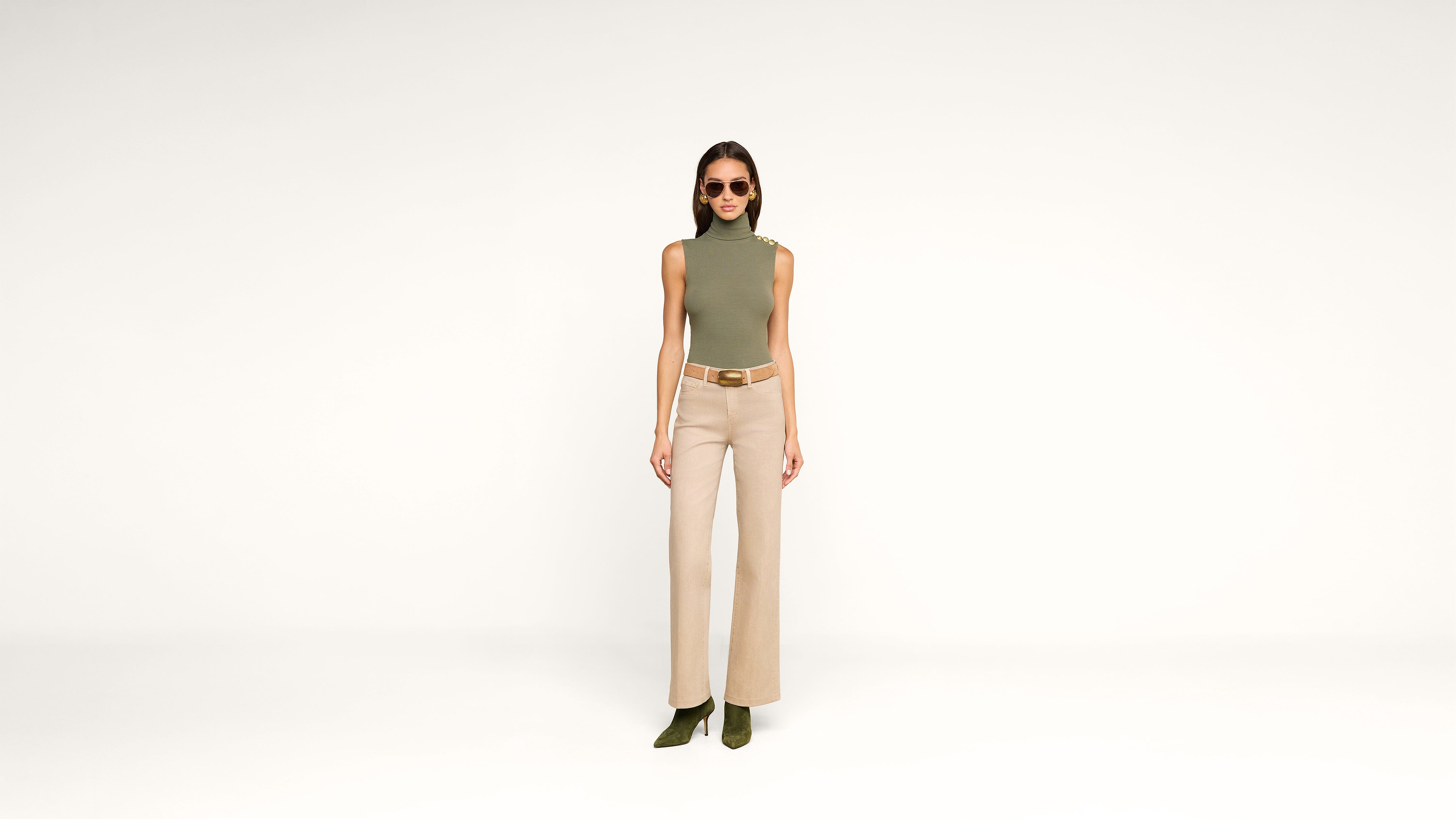 A woman wearing sunglasses, a sleeveless green turtleneck, beige high-waisted pants, and green heels stands against a plain white background.