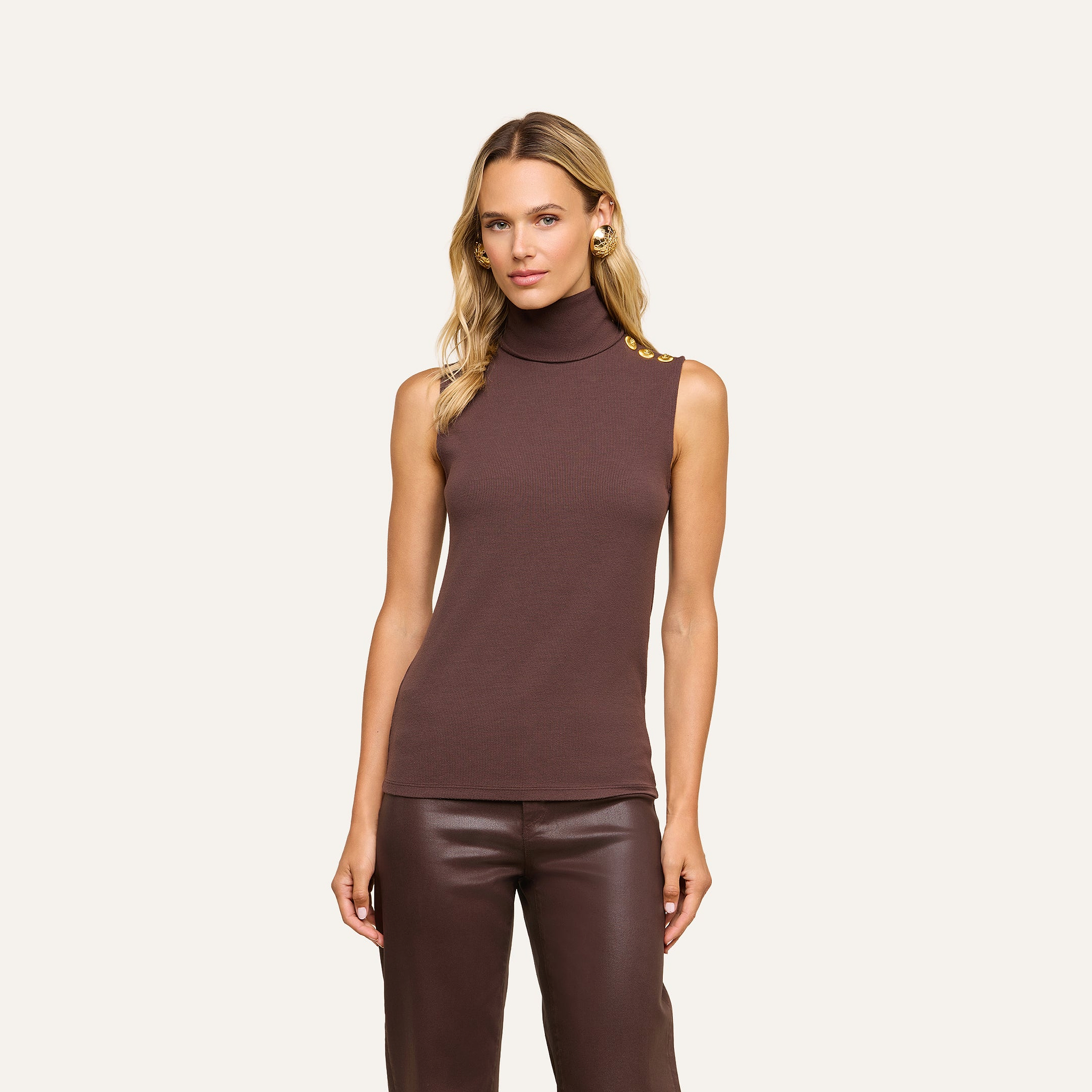 A woman stands facing forward against a beige background, wearing the Ceci Sleeveless Turtleneck—a slim-fit brown turtleneck crafted from stretch rib knit with gold buttons on the shoulder—paired with brown leather pants.