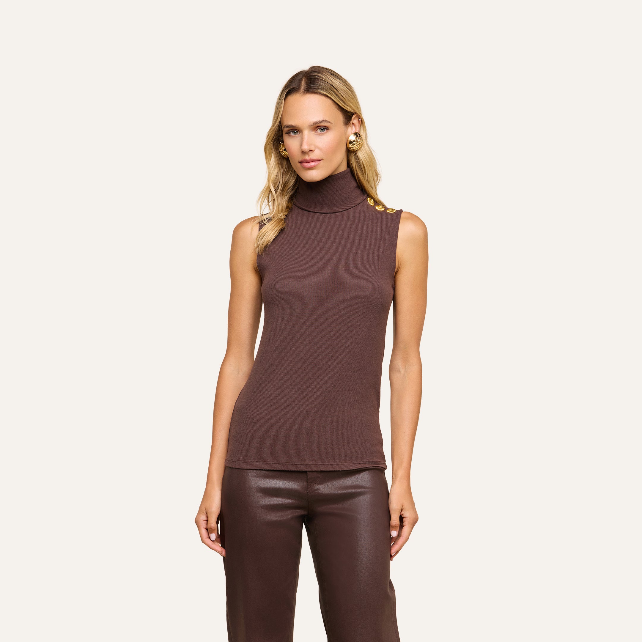 A woman stands facing forward against a beige background, wearing the Ceci Sleeveless Turtleneck—a slim-fit brown turtleneck crafted from stretch rib knit with gold buttons on the shoulder—paired with brown leather pants.