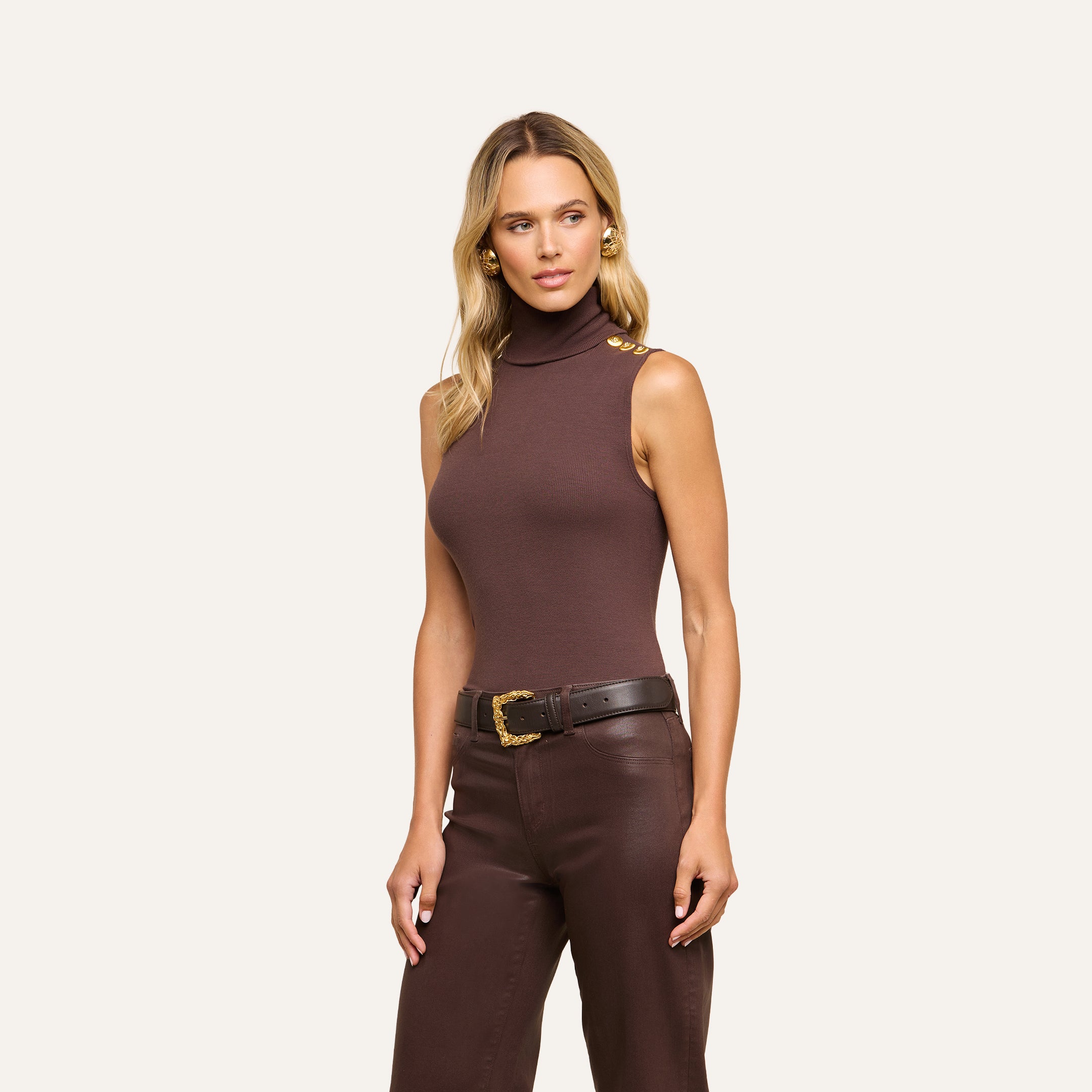 A woman with long blonde hair wears a slim-fit Ceci Sleeveless Turtleneck in brown, crafted from stretch rib knit with gold shoulder buttons, paired with brown pants and a black belt featuring a gold buckle, standing against a neutral background.