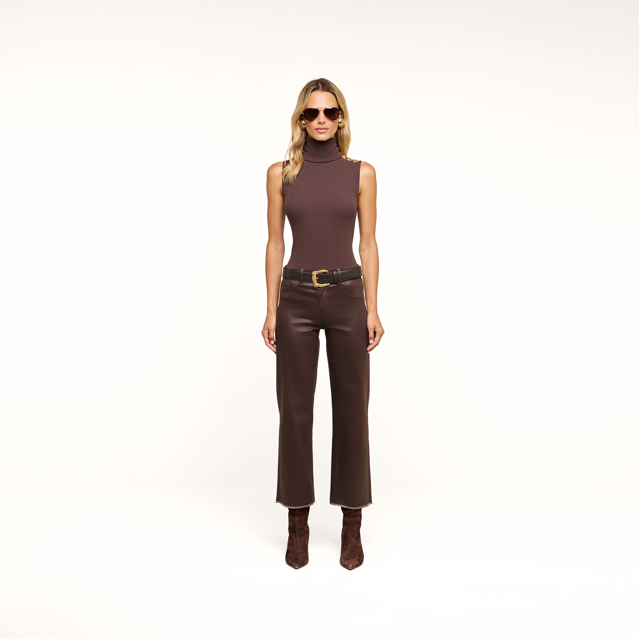 Woman wearing sunglasses, a Ceci Sleeveless Turtleneck in stretch rib knit brown, a brown belt, slim-fit high-waisted pants, and brown boots, standing against a plain beige background.