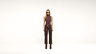A woman stands against a plain white background wearing sunglasses, a sleeveless brown top, brown pants, and brown boots.