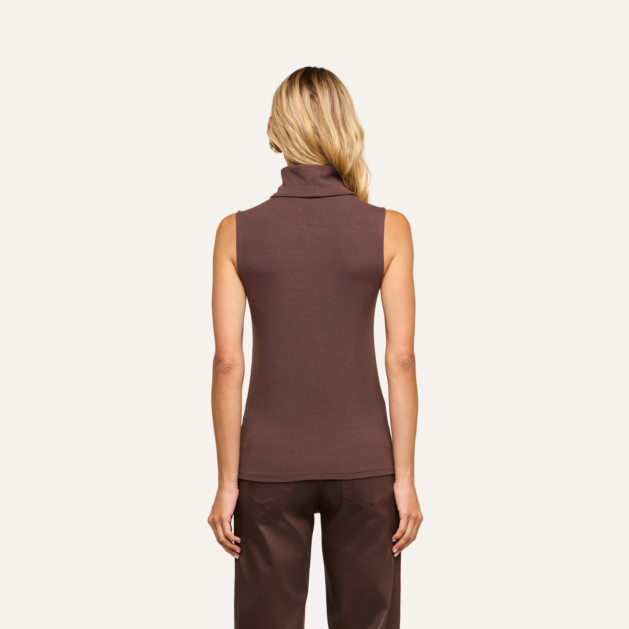 A woman with long blonde hair is shown from the back, wearing a slim-fit, sleeveless brown Ceci Sleeveless Turtleneck crafted from stretch rib knit, paired with dark brown pants, standing against a plain beige background.
