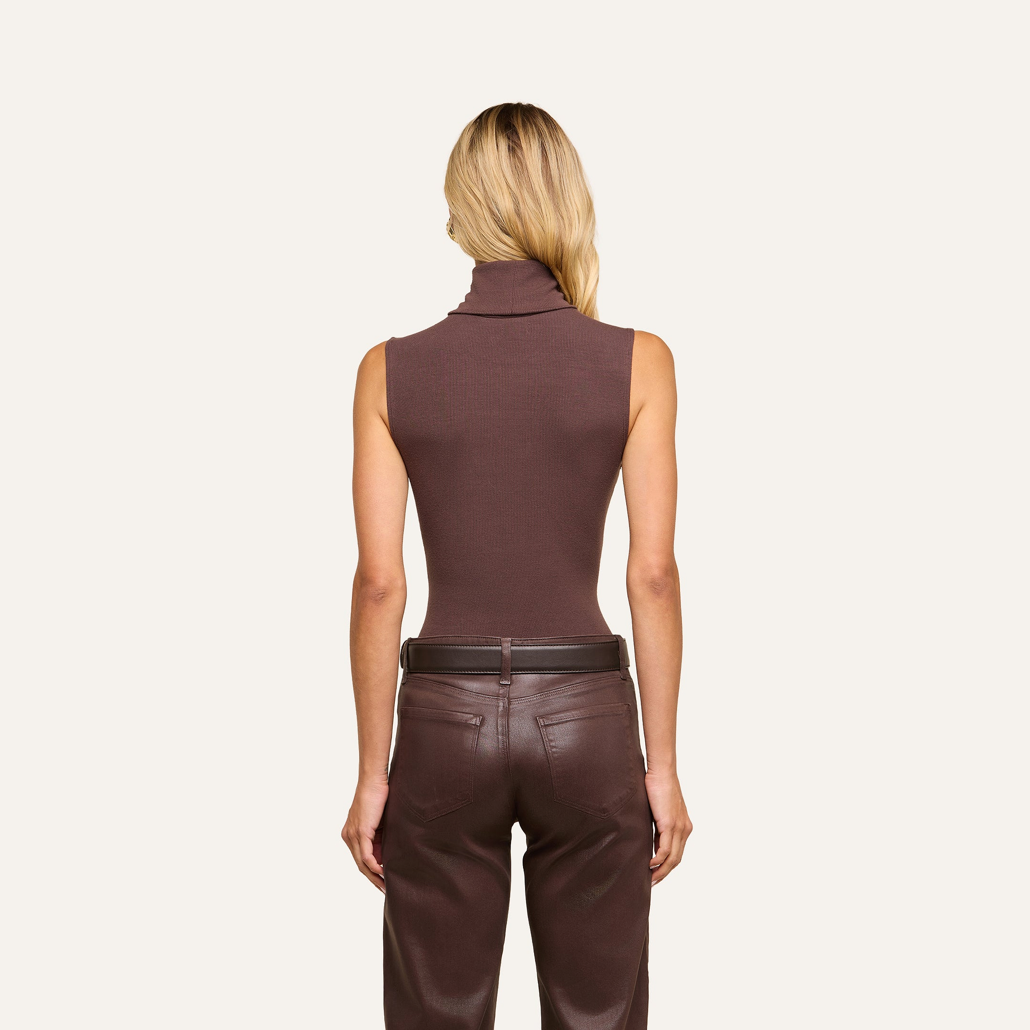 A person with long blonde hair, wearing a slim-fit brown Ceci Sleeveless Turtleneck in stretch rib knit and brown leather pants, stands facing away from the camera against a beige background.