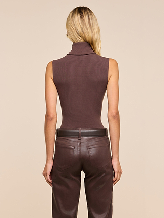 A person with long blonde hair, wearing a slim-fit brown Ceci Sleeveless Turtleneck in stretch rib knit and brown leather pants, stands facing away from the camera against a beige background.