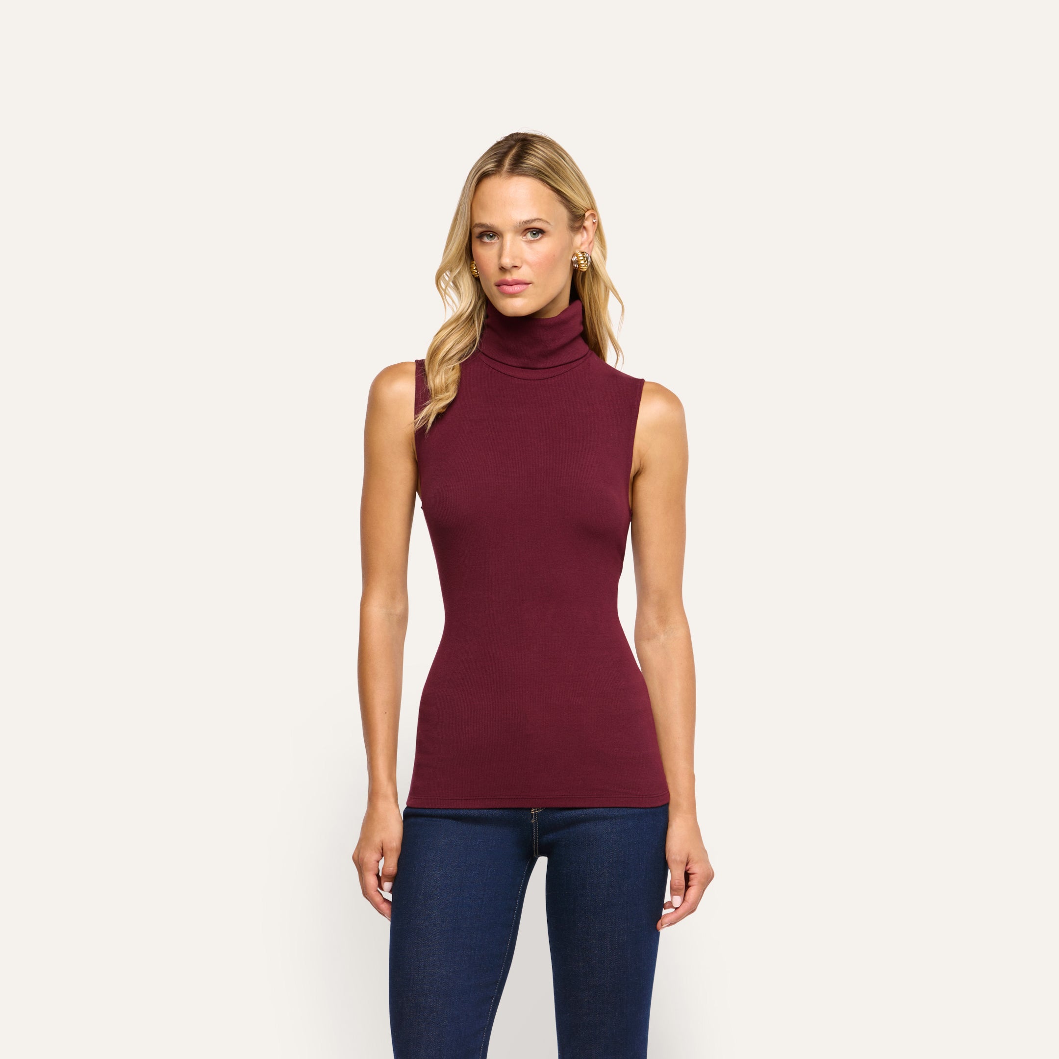 A woman wearing a Ceci Sleeveless Turtleneck in burgundy stretch rib knit with blue jeans.
