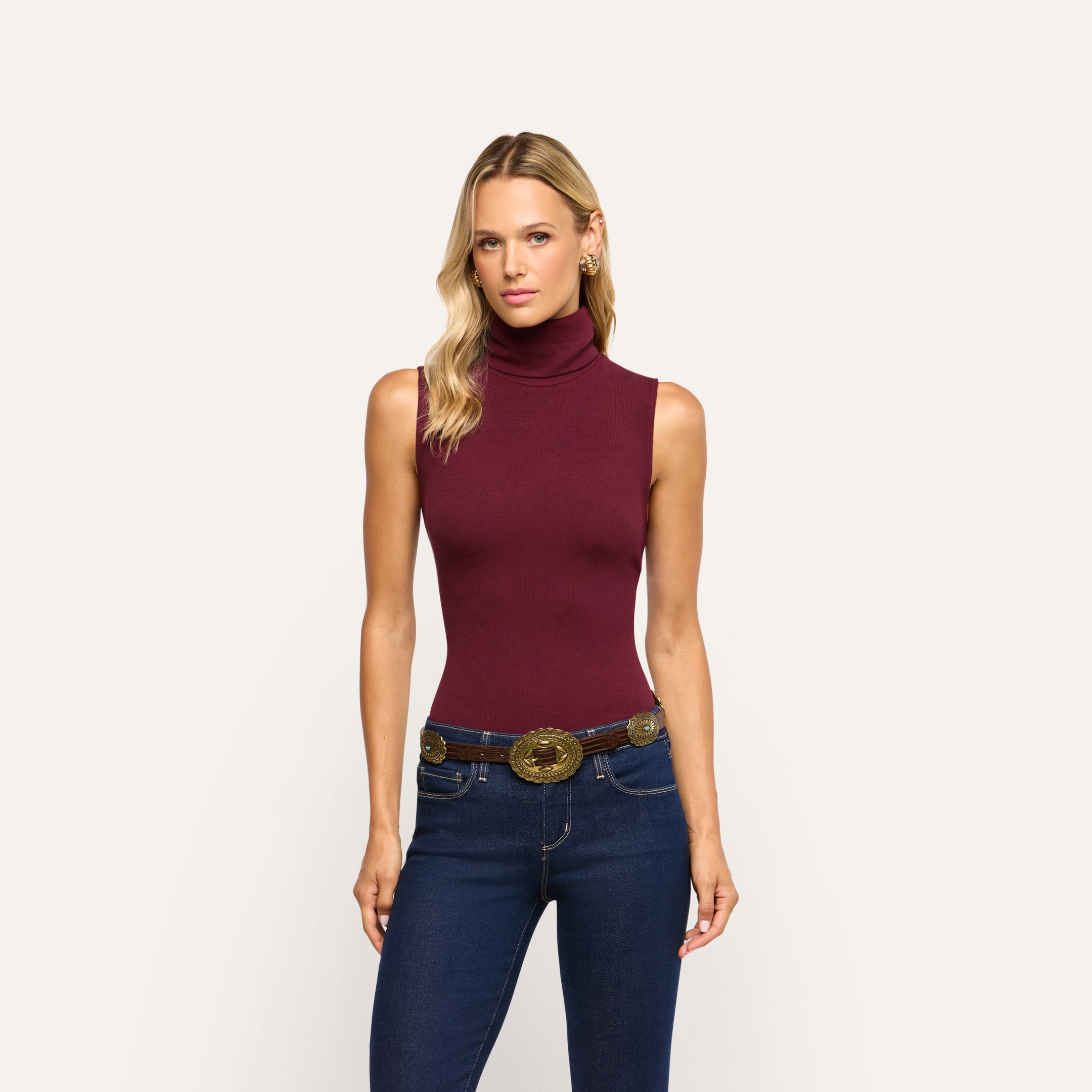 A woman wearing a burgundy Ceci Sleeveless Turtleneck tucked into dark wash blue jeans with a decorative gold-toned belt.