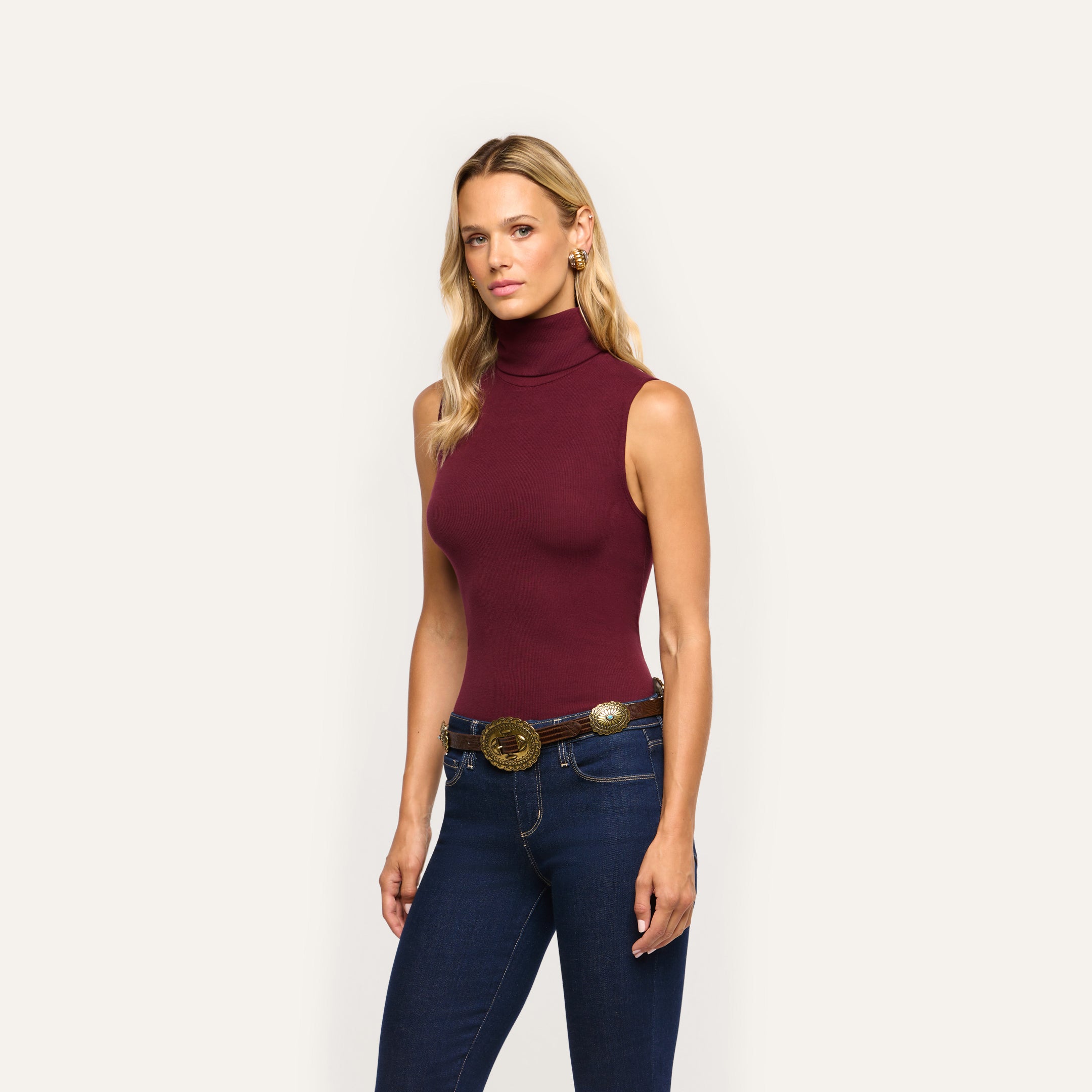A woman wears a slim-fit, sleeveless burgundy Ceci Sleeveless Turtleneck in a stretch rib knit tucked into dark blue jeans with a decorative western-style belt.