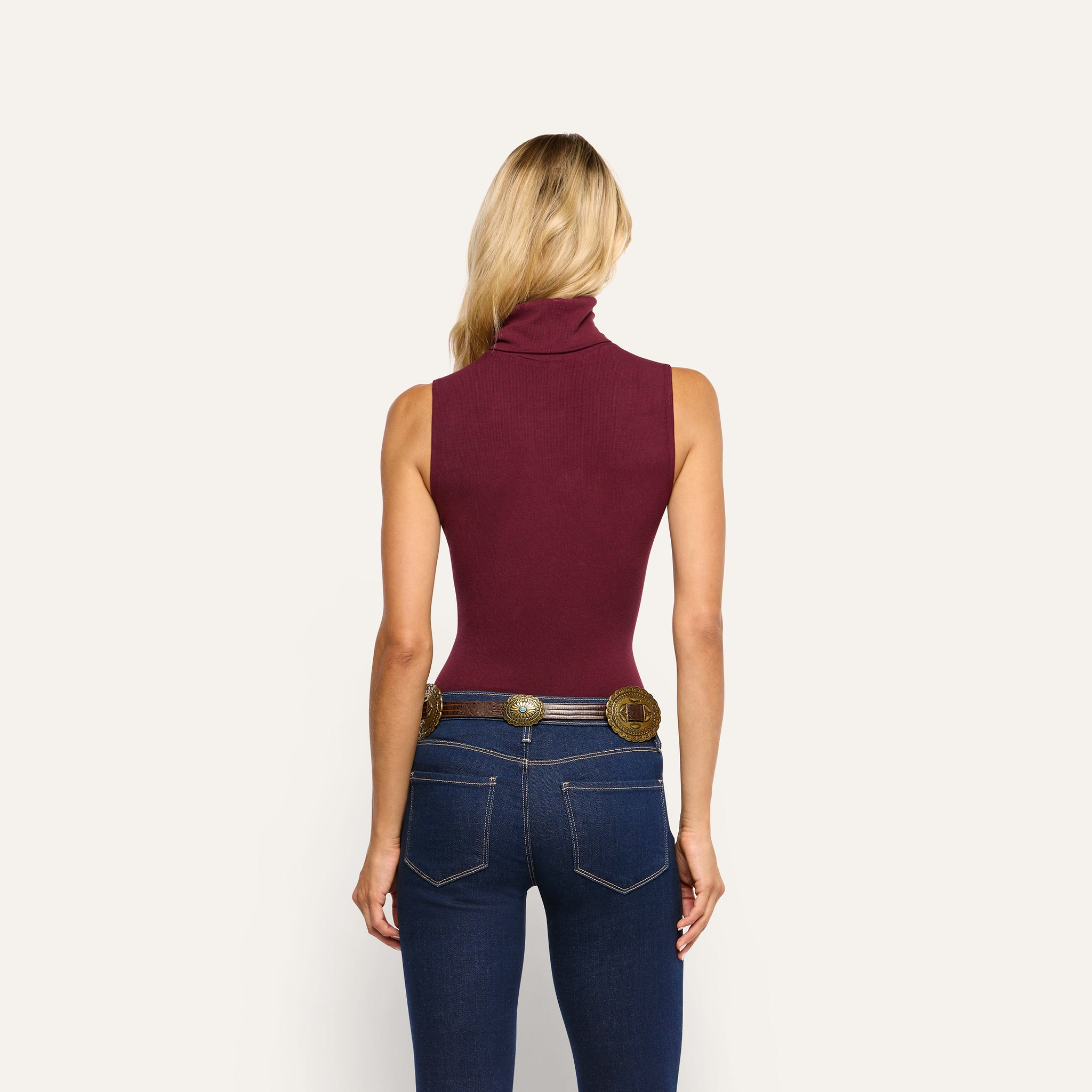 Back view of a person wearing a slim-fit, maroon stretch rib knit Ceci Sleeveless Turtleneck and dark blue jeans with a decorative western-style belt.