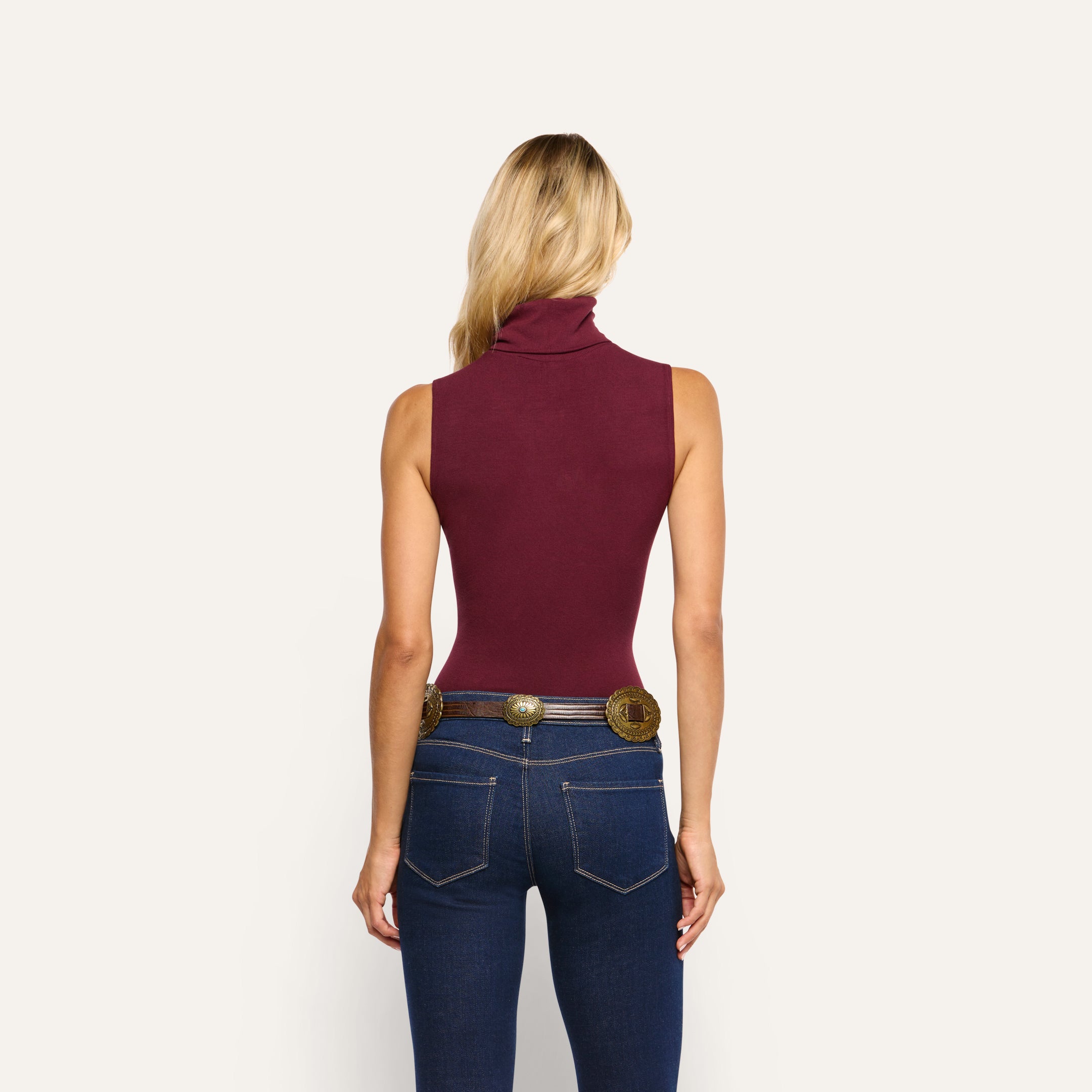 Back view of a person wearing a slim-fit, maroon stretch rib knit Ceci Sleeveless Turtleneck and dark blue jeans with a decorative western-style belt.